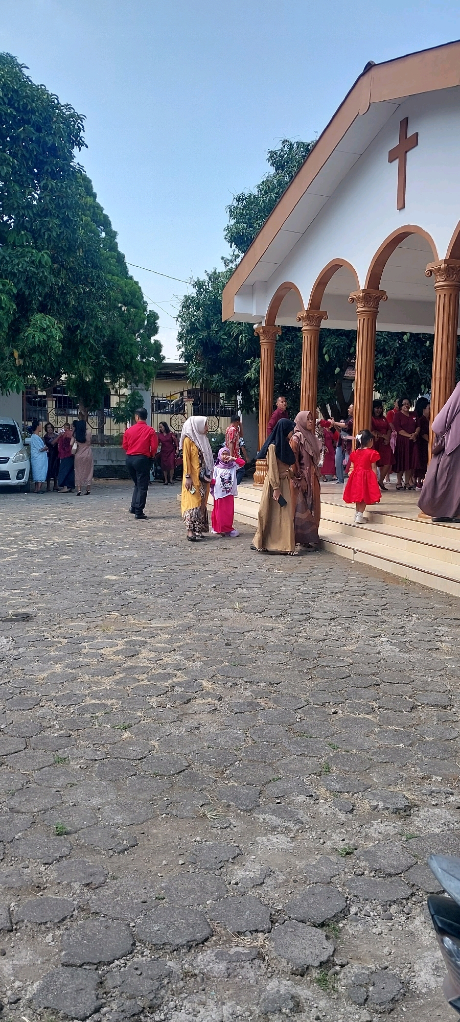 An exterior shot, taken at eye-level, shows the entrance of a church with a cross on its facade. Several people, dressed in traditional clothing, are gathered outside the church. Some are walking towards the entrance, while others are standing and conversing. The ground in front of the church is paved with interlocking hexagonal stones. Tall trees with dense foliage are visible on the left side of the image. The sky is clear and blue. The scene appears to be set in a public area, possibly a church yard. The location is specified as Keling, Indonesia.