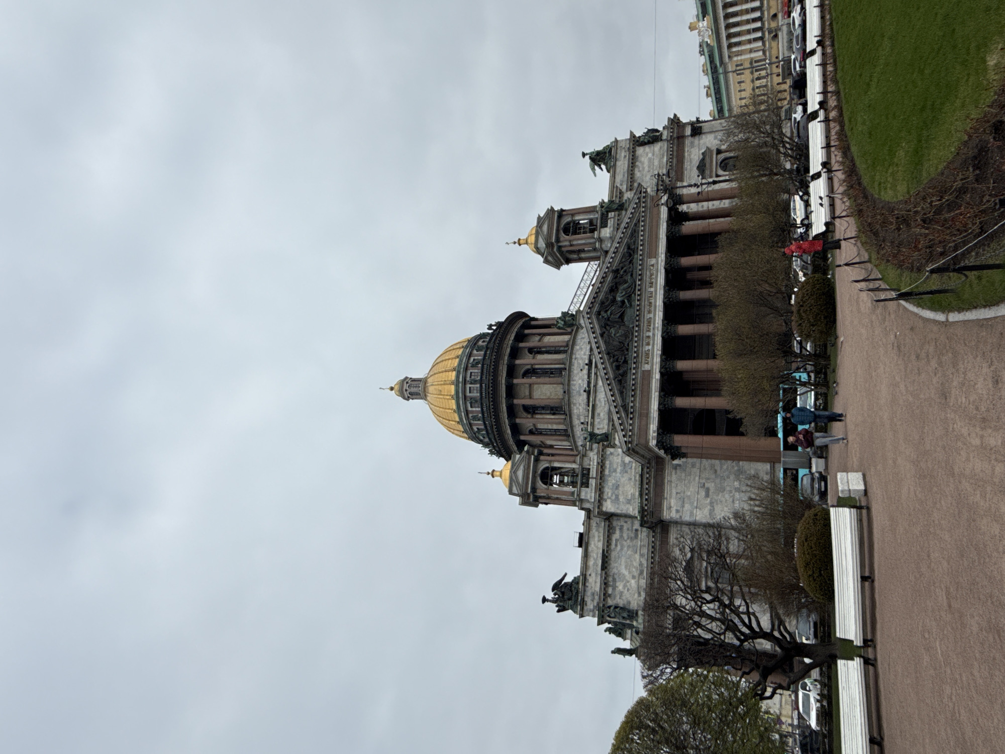 The image captures Saint Isaac's Cathedral in Saint Petersburg, Russia, under an overcast sky. The grand neoclassical structure dominates the left side of the frame, showcasing its massive golden main dome and several smaller golden cupolas. The cathedral's facade is built from light-colored stone, characterized by numerous tall columns and intricate sculptural elements, including figures on the pediments and around the base of the dome.
In the mid-ground, a wide, unpaved pathway runs along the building, separating it from a strip of green grass and several deciduous trees with bare branches, suggesting early spring or late autumn. Multiple white slatted benches are placed beside the path. A few people are visible; one person in a distinctive red jacket stands near a bush, while others, including a pair, are walking further down the path towards the right, near parked cars and what looks like a public transport stop.
The sky is a uniform, pale grey, indicating a heavily clouded day with no direct sunlight, creating a muted and possibly cool atmosphere. Some Cyrillic text is visible inscribed on the frieze below the pediment on the cathedral's facade, though it is not clearly legible from this distance. The scene conveys a sense of historical majesty and urban tranquility on a typical cloudy day.