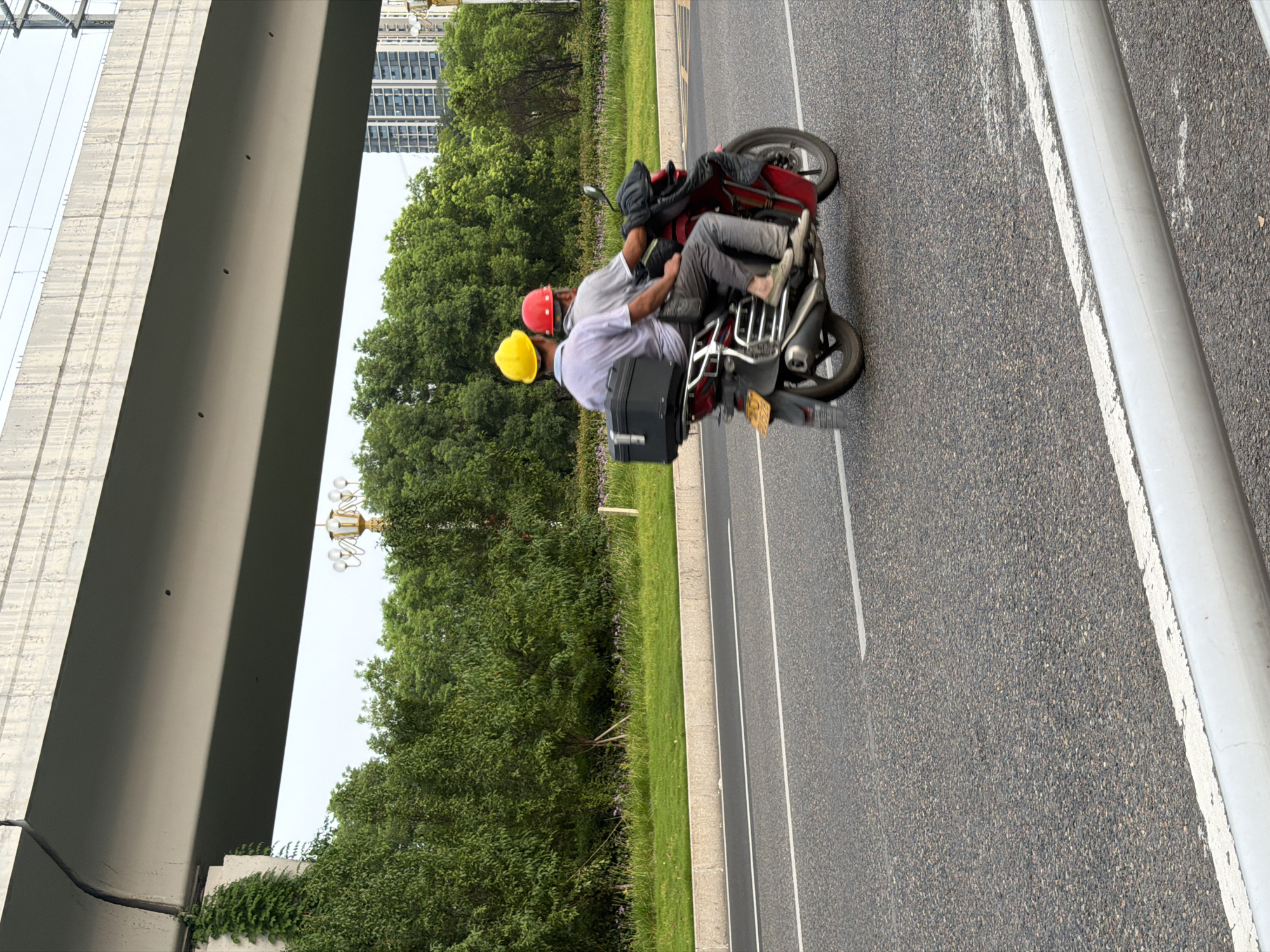 Two individuals, both wearing hard hats, are riding on a red motorcycle on an asphalt road. The person in the front is wearing a red hard hat and a grey shirt, while the person in the back is wearing a yellow hard hat and a light-colored shirt. They are positioned side-by-side, with the passenger in the rear. A black storage box is attached to the back of the motorcycle behind the passenger. The motorcycle is moving from left to right in the frame. In the background, there are lush green trees and a tall building visible. An elevated concrete structure, likely an overpass or bridge, spans across the upper portion of the image. The scene appears to be in an urban or suburban area of Jiangjin, China.