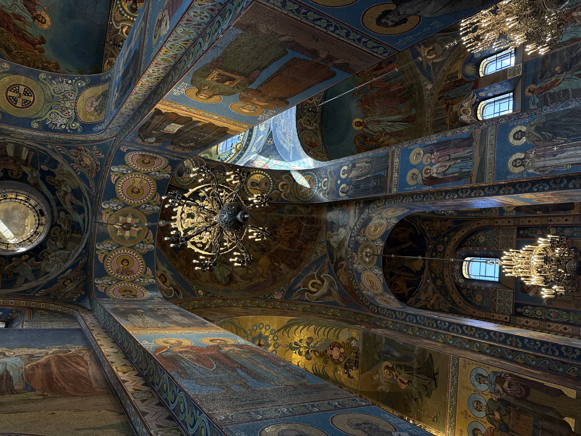 The image depicts the interior of the Church of the Savior on Spilled Blood in Saint Petersburg, Russia. The ornate ceiling and walls are covered in intricate mosaics, featuring religious figures and patterns in vibrant blues, golds, and reds. A large, multi-tiered chandelier hangs from the center of the ceiling, with numerous lit bulbs casting a warm glow. Another chandelier is visible in the lower right corner of the image. Sunlight streams through arched windows, illuminating sections of the mosaics and creating dramatic light and shadow effects. The architecture is characterized by soaring arches and domed structures, all elaborately decorated. There are no people visible in the frame. The overall impression is one of grandeur and artistic opulence.