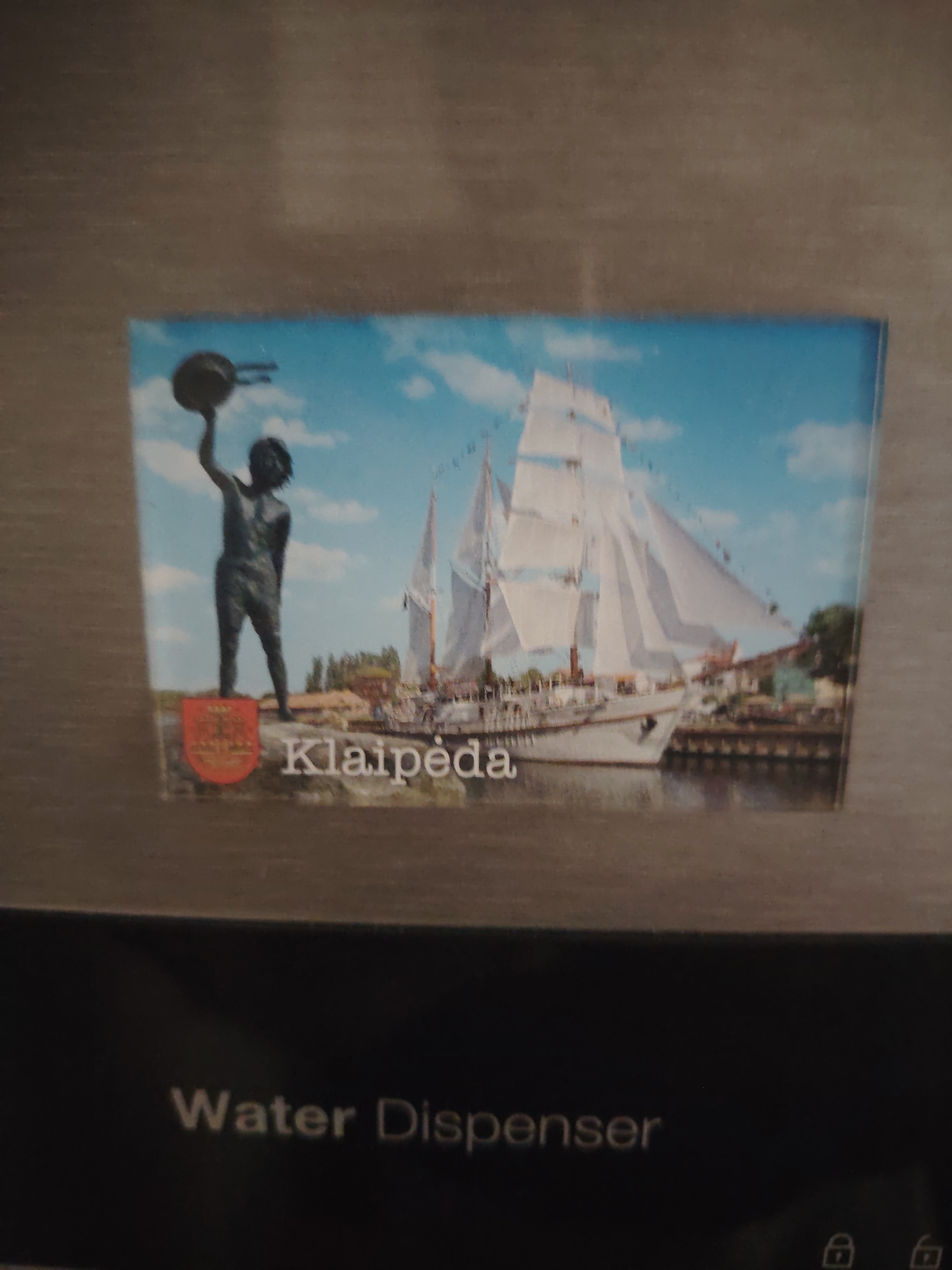 The image displays a rectangular magnet or postcard adhered to a light grey, brushed metallic surface. The magnet features a scenic depiction, primarily split between a statue and a ship under a partly cloudy blue sky. On the left side of the magnet, a dark-colored statue of a young person, possibly a boy or a man, stands with their left arm raised, holding a round object resembling a pot or a cap. Below the statue is a red heraldic shield with yellow detailing. On the right side, a large white sailing ship with multiple masts and sails is depicted on calm water, likely in a port or harbor, with trees and buildings visible along the distant shore. The word "Klaipėda" is prominently displayed in white letters at the bottom center of the magnet. Below the magnet, on a dark, matte surface, the white text "Water Dispenser" is horizontally aligned. Further to the bottom right, faint icons resembling lock and unlock symbols are visible. The scene depicted on the magnet is of Klaipėda, a city in Lithuania, contrary to a location in Huntingdonshire, United Kingdom.