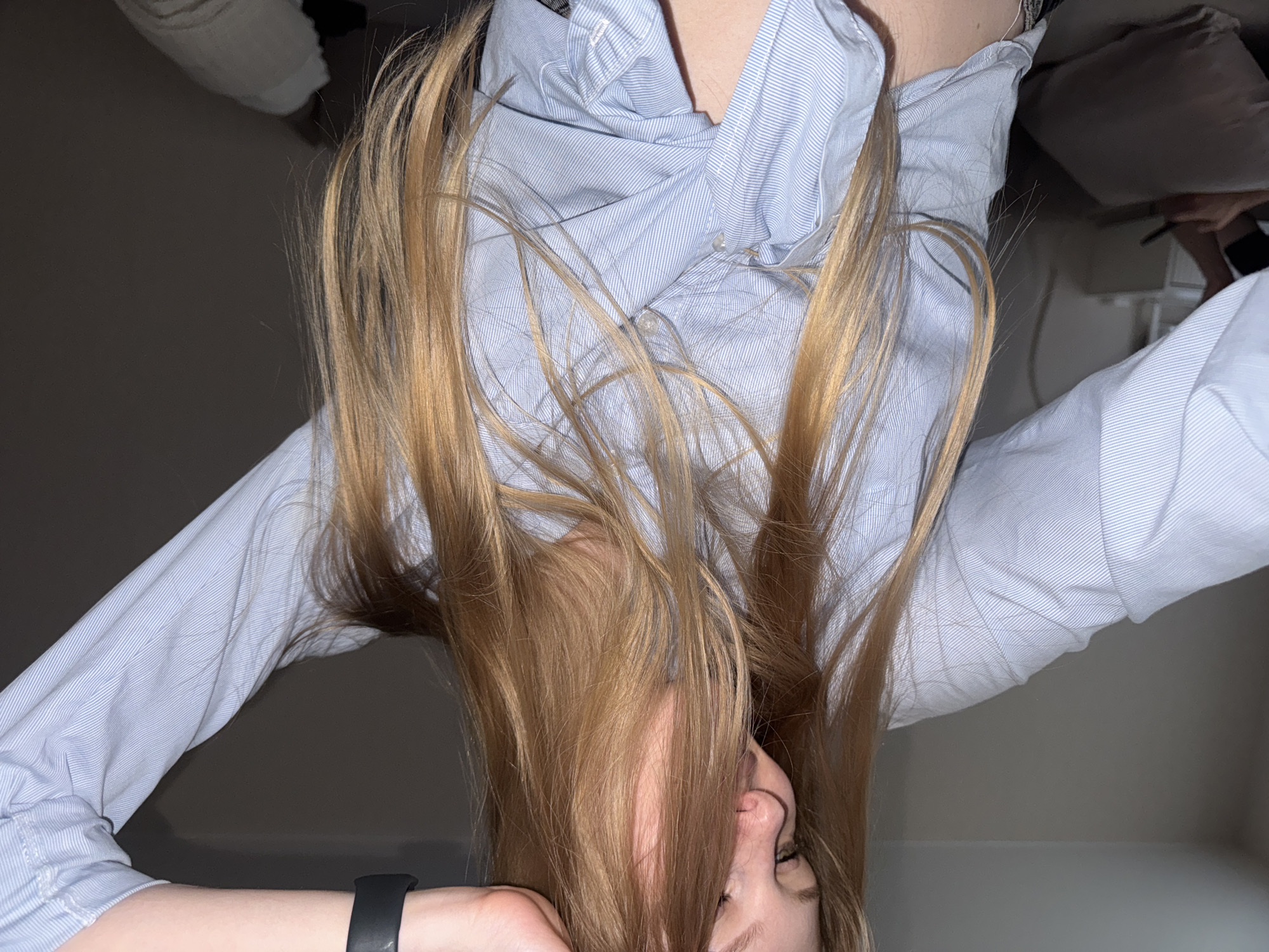 A young woman with long, blonde hair is shown from the chest up, wearing a light blue button-down shirt tied at the waist. She is looking downwards, her hand raised to her head. The background is a dimly lit room with a gray wall and hints of furniture. No specific location within the city of Perm, Russia can be identified from the image.