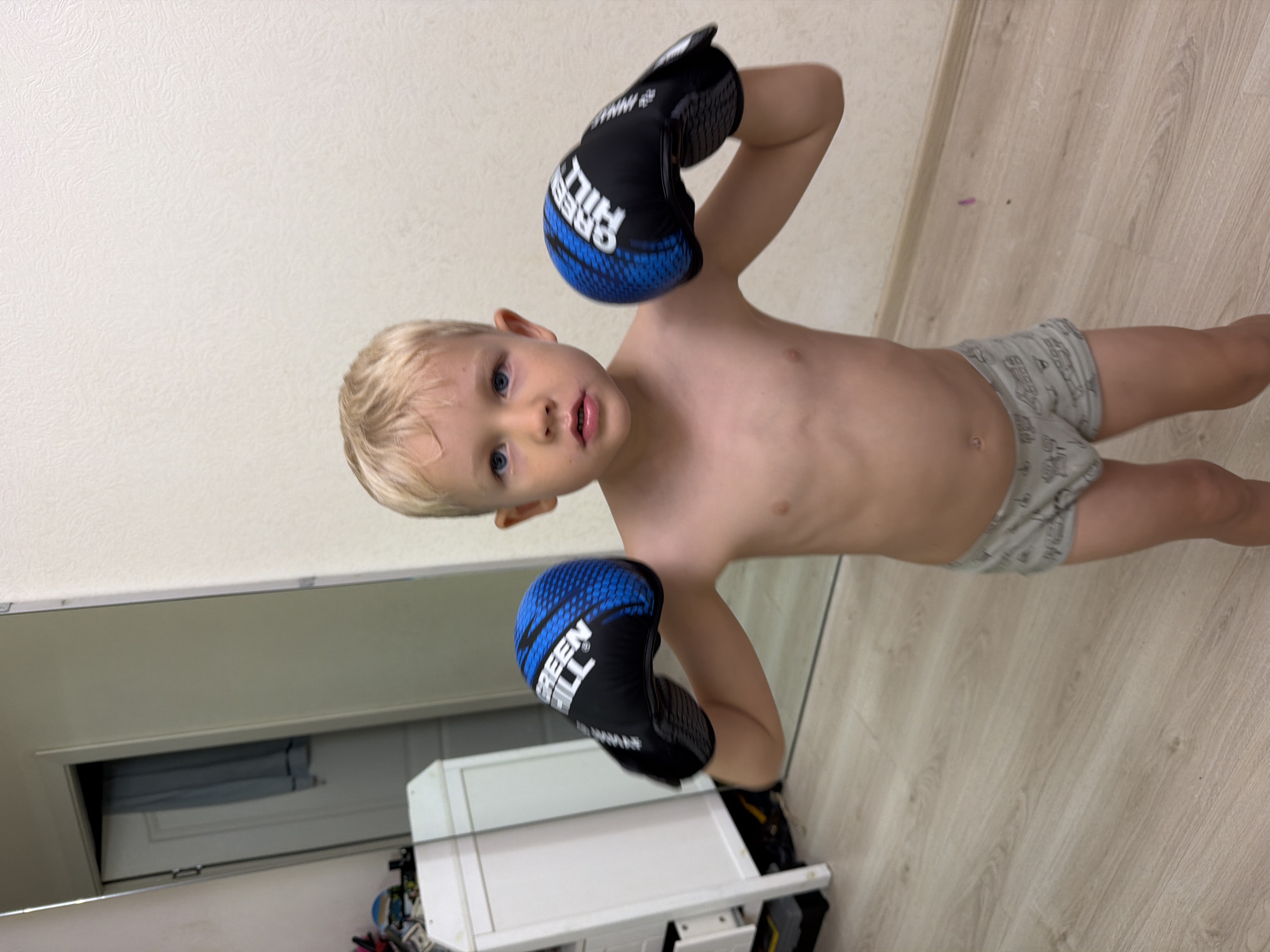 A young, fair-skinned child with blond hair is posing indoors, wearing boxing gloves and underwear. The gloves are black with blue accents and the brand "Green Hill" is visible. The child's skin is pale. The child's underwear is gray with illustrations on them. The background includes a white wall, light-colored wooden flooring, a partially visible mirror, and a white storage unit with miscellaneous items beneath. The location is potentially a home or residential space, with no indication of being in Sochi, Russia. The orientation of the image suggests it was taken with a phone, with the subject rotated.
