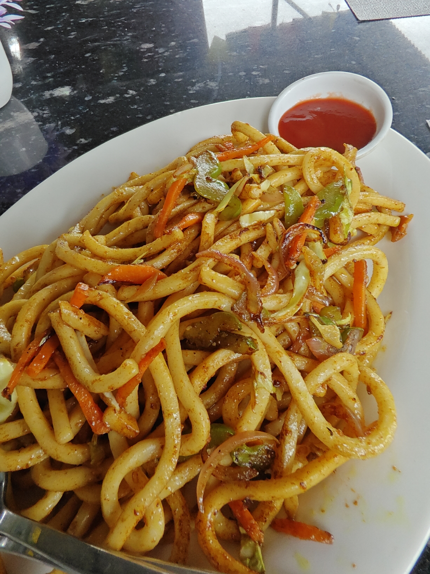 A plate of noodles with vegetables and a small dish of ketchup sits on a dark, speckled countertop in Pokhara, Nepal. The noodles appear to be thick and stir-fried with julienned carrots, onions, and green bell peppers. A white plate holds the generous portion of noodles, and a small white dish containing a red sauce, likely ketchup, is positioned in the upper right corner of the plate. The background shows a dark, reflective surface, possibly a table or countertop, with subtle patterns. The lighting suggests an indoor setting, possibly a restaurant. There are no people visible in the image.