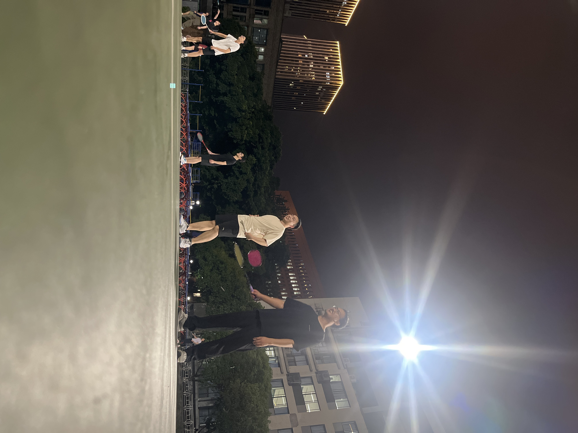 In Yuzhong District, China, under the illumination of a bright overhead light, five individuals are engaged in a game of badminton on an outdoor court at night. The court is a flat, green surface. In the foreground, the legs and lower bodies of two individuals are visible, one in black pants and the other in black shorts. Further back, four men are positioned on the court, each holding a badminton racket. One man in a white t-shirt and black shorts is standing, facing left, while another in a tan t-shirt and black shorts is also standing. A third man, wearing a black t-shirt and black shorts, is in mid-action, holding a red racket. The fourth man, in a black t-shirt and black pants, is also holding a racket and looking to the right. In the background, two modern, tall buildings with illuminated facades rise against the dark night sky, characteristic of a city environment. Trees line the perimeter of the court, and a blue net is visible. The overall scene depicts a casual recreational activity taking place in an urban setting at night.