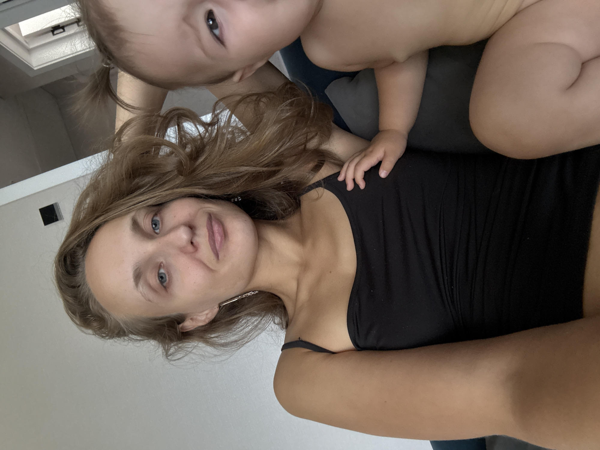 In this close-up, vertically oriented image, a woman and a baby are shown in a close embrace. The woman, with light brown hair that cascades around her shoulders, looks directly at the camera with a gentle smile and blue eyes. She is wearing a black tank top. A silver earring is visible on her right ear. The baby, positioned above and slightly behind the woman, has light brown hair tied in a small ponytail and is looking upwards and to the side. The baby's hand rests on the woman's shoulder, indicating a moment of affection.
The background is softly lit, suggesting an indoor setting. A skylight is partially visible in the upper left corner of the image, hinting at a room with natural light. The overall mood of the image is intimate and warm, capturing a tender moment between mother and child. The specific location is identified as Podshivalovo, Russia. No text is visible in the image.