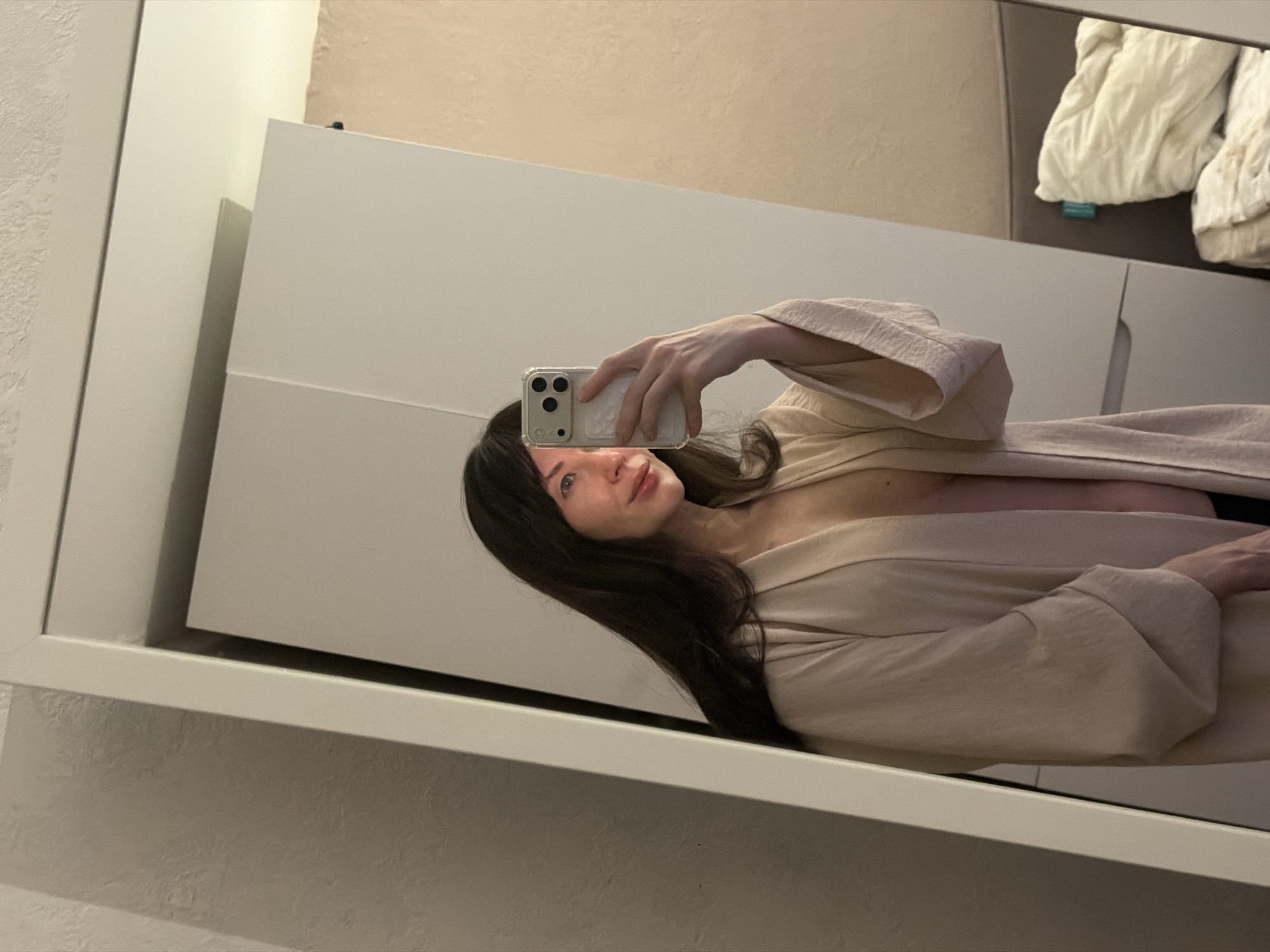 A woman with dark hair takes a selfie in a mirror. She is wearing a light-colored robe or wrap. The background includes a white cabinet or closet and a wall of light beige or tan color. White bedding is visible in the upper right corner of the image. The location is difficult to determine from the image, so I can't identify it.