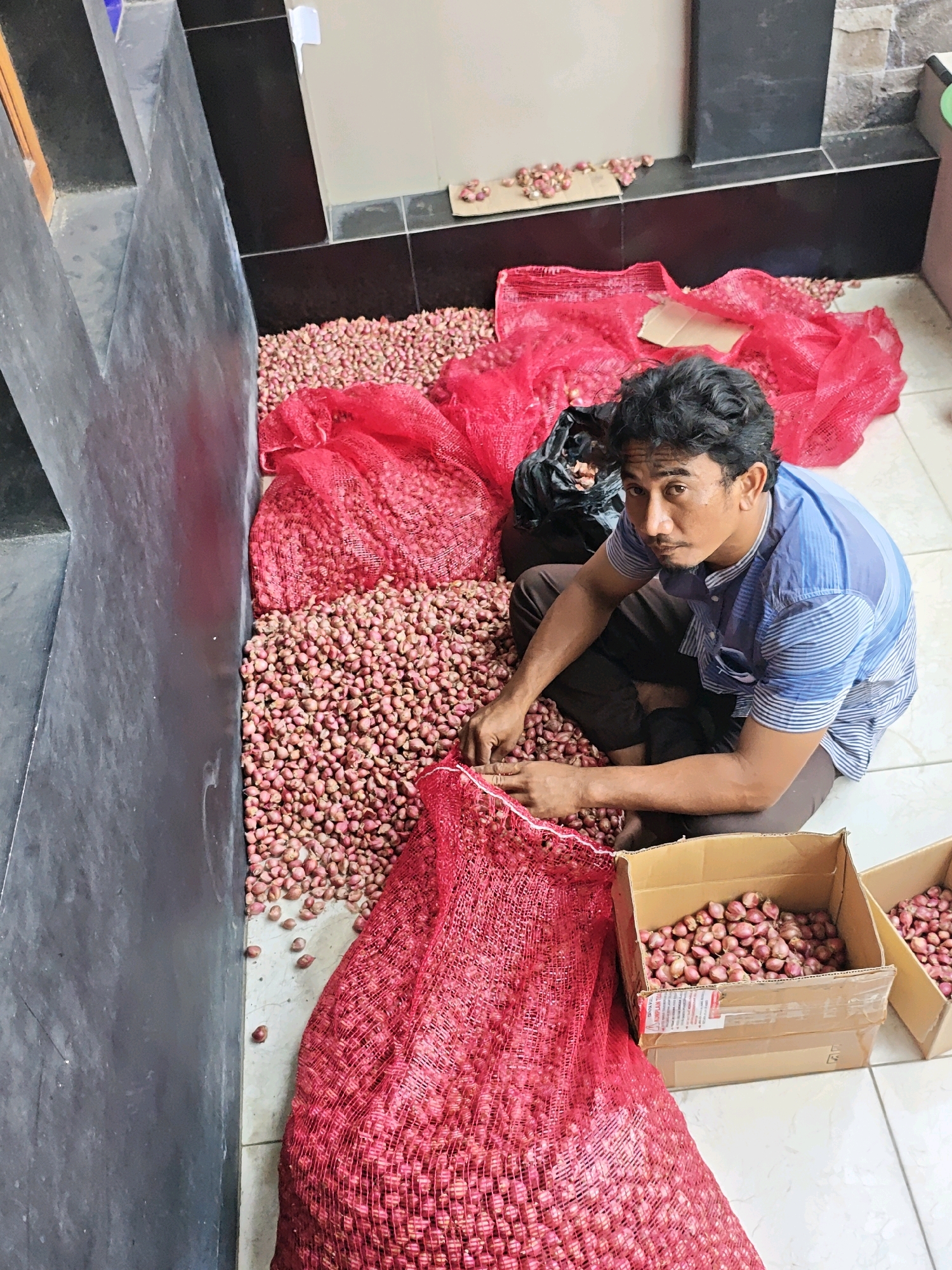 A middle-aged Indonesian man with dark, curly hair and a mustache is sitting on the floor in what appears to be a storage area, surrounded by large quantities of shallots. He is wearing a blue and white striped short-sleeved shirt and brown pants. The man is crouched amidst piles of shallots, with some contained in red mesh bags and others spread loosely on the floor. Two cardboard boxes filled with shallots are placed to his right. The floor is tiled with light-colored squares. In the background, dark gray tiled walls suggest an indoor setting. Scattered shallots are also visible on cardboard pieces on the floor near the wall. The scene suggests a wholesale or retail operation for shallots, possibly in Ngasem, Indonesia, given the context.
