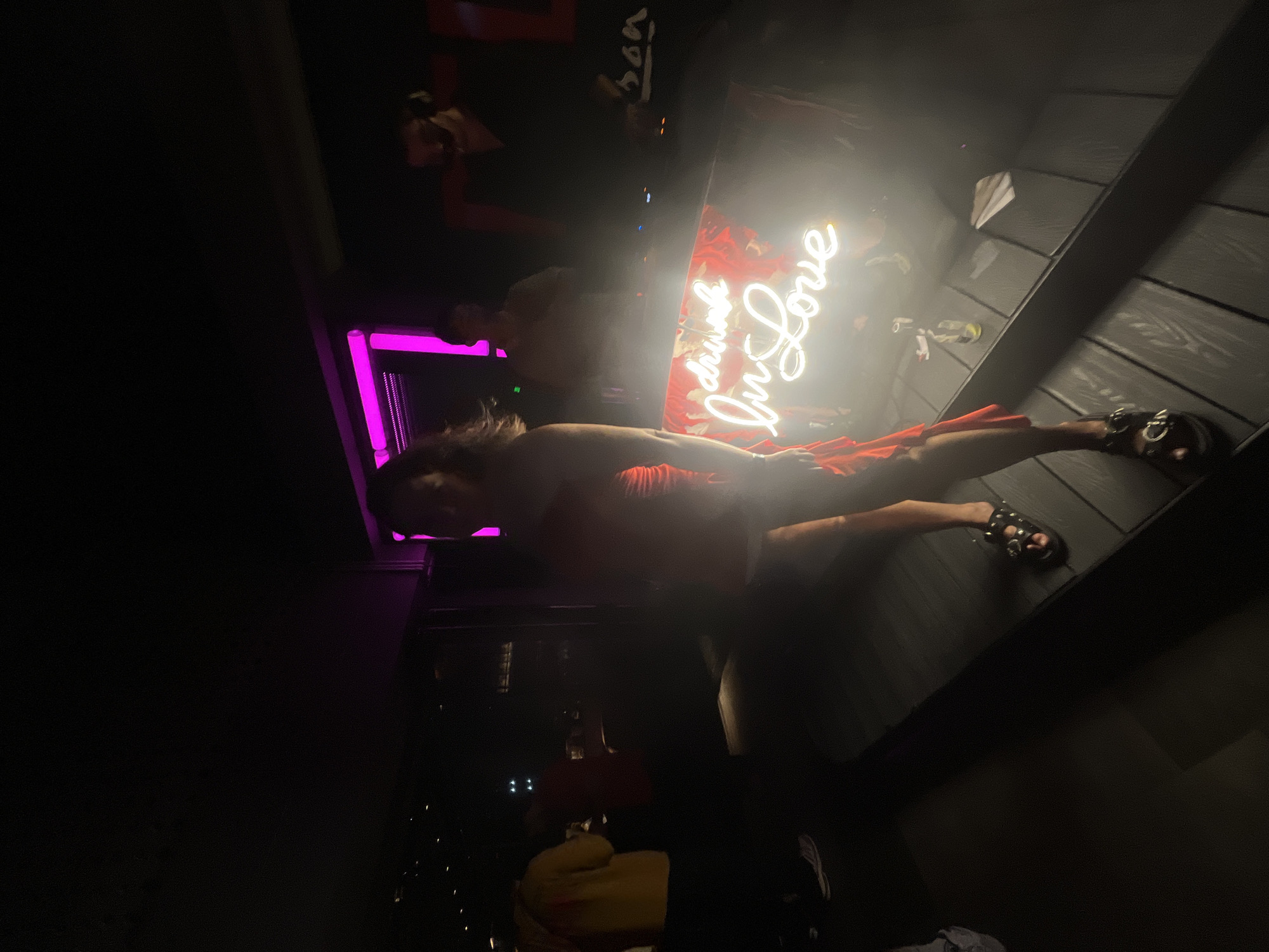 In a dimly lit nightclub in Da Nang, Vietnam, a woman in a striking red dress stands center frame. Her dress, with its ruffled detailing and strapless design, accentuates her figure. She wears black studded sandals, and her stance suggests a moment of pause or a dance move captured.
Behind her, a neon sign glows, spelling out "drink in Love" in cursive script. The sign is affixed to a backdrop of a vibrant floral pattern. To the right of the woman, a bar or DJ booth is visible, with indistinct figures of people working or enjoying themselves. Further back, in the background, other patrons are present, some in yellow and red clothing, contributing to the lively atmosphere of the club. The overall setting is dark, with purple and pink lighting creating a vibrant, nocturnal ambiance.