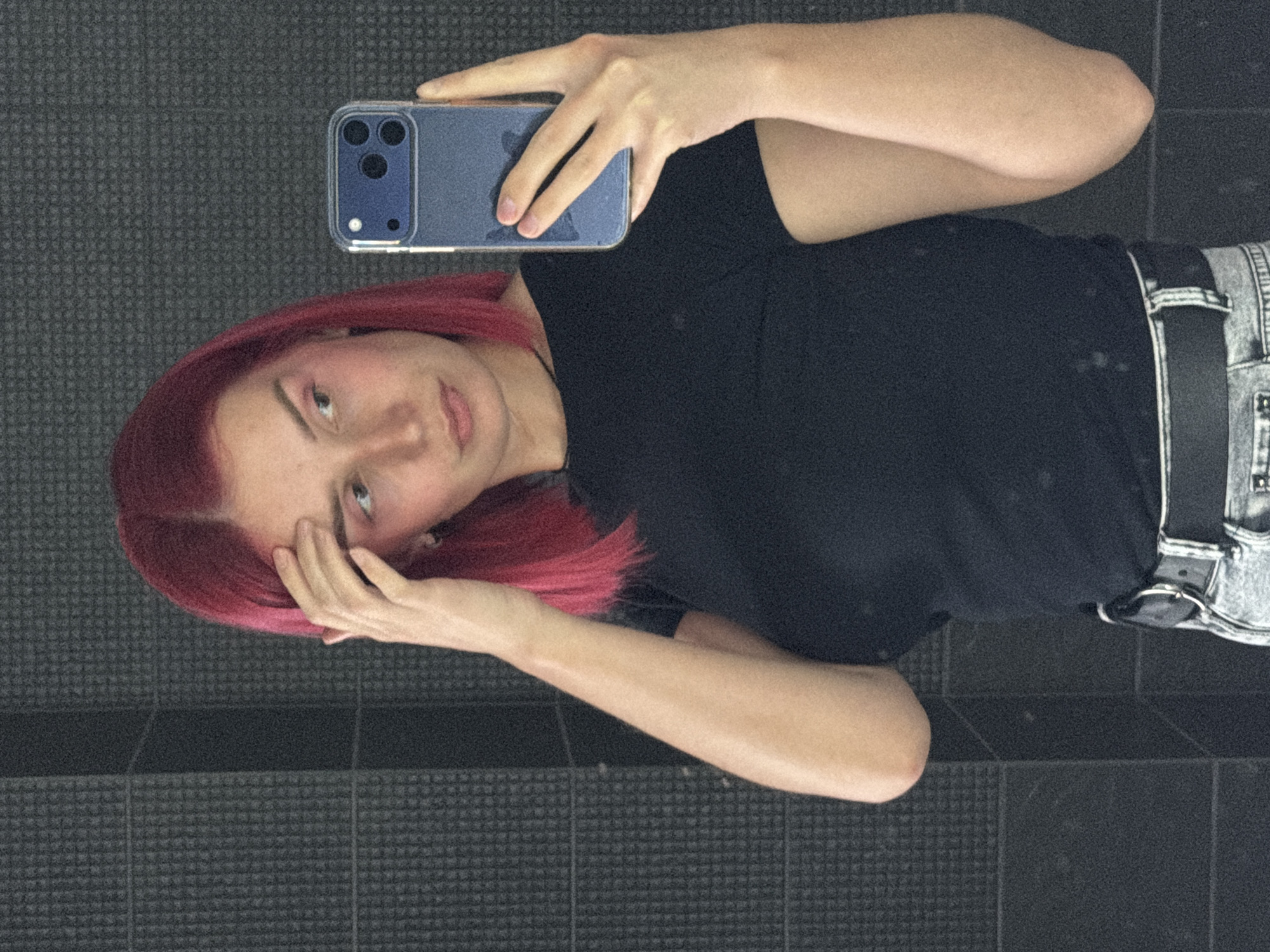An indoor selfie captured in Chaikovsky, Russia, shows a young individual with reddish-pink hair reflected in a dark, glossy surface, likely a mirror or highly polished floor/wall. The person has fair skin and light-colored eyes, looking towards the camera with a neutral to slightly pensive expression. They are wearing a plain black t-shirt, light-wash denim jeans, and a black belt with a silver buckle.
The individual holds a blue smartphone, possibly an iPhone with a clear case showcasing its triple camera setup, in their left hand to take the photo. Their right hand is raised, lightly touching their hair near their face. The background, which is the actual surface behind the reflection, consists of dark, square-patterned tiles, suggesting a modern interior space such as a bathroom, gym, or public area. The lighting is soft and even, typical of indoor artificial illumination. No discernible text is visible on the person's clothing or the surrounding environment.