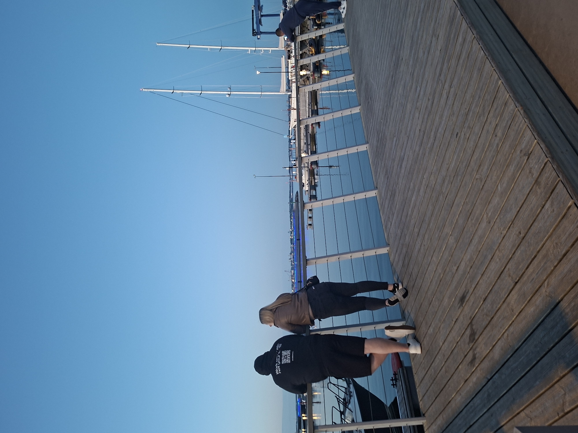 The image depicts a marina in Tallinn, Estonia, at dusk or early evening. A wooden boardwalk extends from the foreground towards the mid-ground, bordered by a modern railing with white vertical posts and horizontal dark wires.
On the left side of the boardwalk, two individuals lean against the railing, facing away from the viewer and looking towards the water and distant boats. The person on the left wears a black hooded sweatshirt with white text on the back that reads "Work on music not drugs" above a QR code-like graphic and "BASEBALL." They also wear black shorts and white sneakers. Next to them, a person with blonde hair wears a brown jacket, dark pants, and dark sneakers with white soles.
Further down the boardwalk to the right, a third person in a dark blue long-sleeved top and matching pants, with white sneakers, leans forward over the railing, observing the scene.
The calm water, which is a dark blue, reflects subtle lights. Several boats are docked in the marina. To the far left, a dark-hulled smaller boat is partially visible. In the mid-ground, at least two large white sailboats with tall masts are prominently visible against the sky. Across the water, distant illuminated structures, possibly buildings or cranes, are visible with blue and white lights. The sky is clear, displaying a gradient of light to darker blue indicative of twilight.