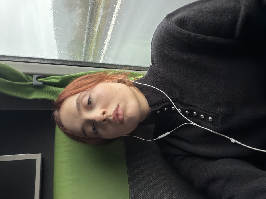 The scene depicts a young adult female seated inside a moving vehicle in Moineville, France. She has reddish-brown hair and a light complexion, looking directly at the camera with a neutral expression. She is wearing a black, long-sleeved knitted sweater featuring a high collar secured by five visible silver snap buttons down the front placket. White wired earphones are inserted into both ears, with the cable extending down her chest and an inline remote control unit visible. To her left, part of a green fabric seat or headrest is visible, along with a dark rectangular screen or panel. To her right, a green fabric curtain is pulled back by a grey plastic clip, revealing a rain-streaked window. Through the window, a blurred, overcast, and wet exterior environment, possibly a highway with faint white lane markings, is visible, indicating motion and precipitation.