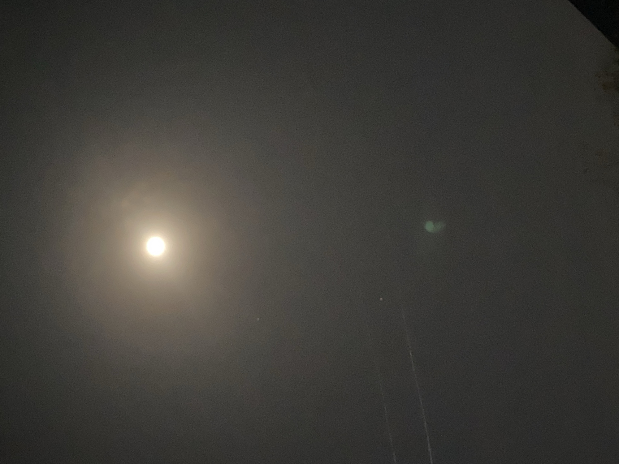 The image depicts a dark, likely nighttime, sky. A bright, circular light source, presumably the moon, is visible towards the left side of the frame. The sky appears overcast or foggy, obscuring the details. Two tall, thin structures, perhaps poles or towers, are vaguely visible towards the right side of the image. A slight green haze appears to the right of the moon. The location of Jos, Nigeria, is not directly evidenced in the image and cannot be confirmed.