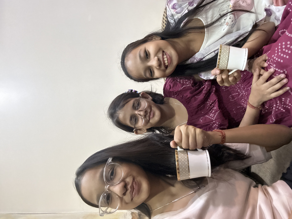 Three young women are angled in the frame, each holding a white mug with a gold design. The top girl wears a light-colored patterned top and smiles widely. The middle girl wears glasses and a dark patterned top, also smiling. The bottom girl, closest to the camera, sports clear-framed glasses and a light pink top. They appear to be indoors.