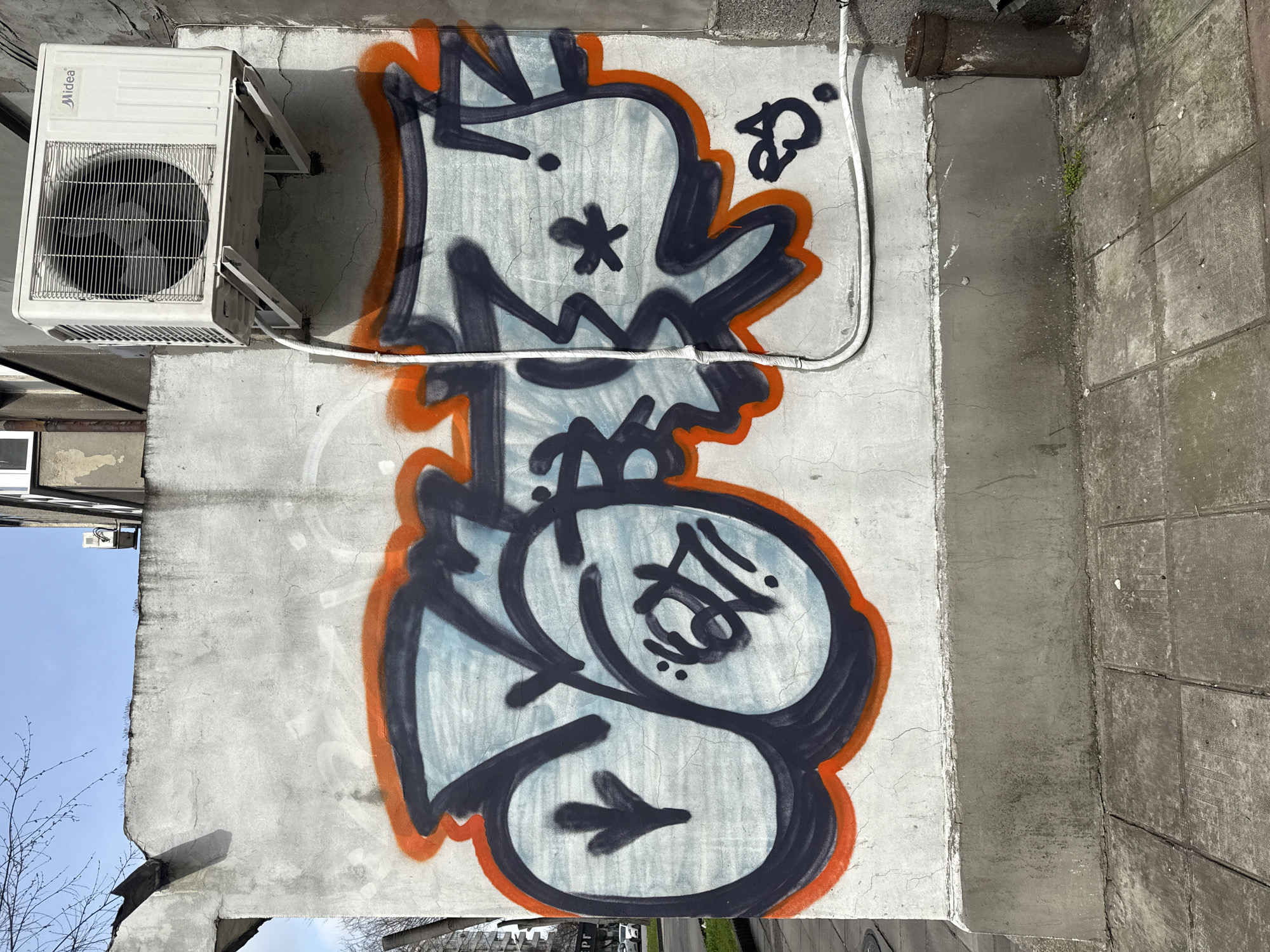 A vertical shot shows a section of a concrete building exterior with graffiti. The graffiti consists of an abstract cartoonish character outlined in orange and filled with light blue. The outlines of the character are thick black, with a star-like shape for an eye and other ambiguous symbols. To the left and above the graffiti is a Midea air conditioning unit. A white cable runs from the unit along the wall to the right of the graffiti. To the right of the wall section with graffiti is a darker concrete section with rectangular patterns. In the upper right corner, a rusty pipe protrudes from the wall. The bottom of the image shows a glimpse of a city street with buildings and trees. The upper left corner shows a blue sky. The image is likely taken in Varna, Bulgaria, based on the characteristics of the buildings and street visible in the background.
