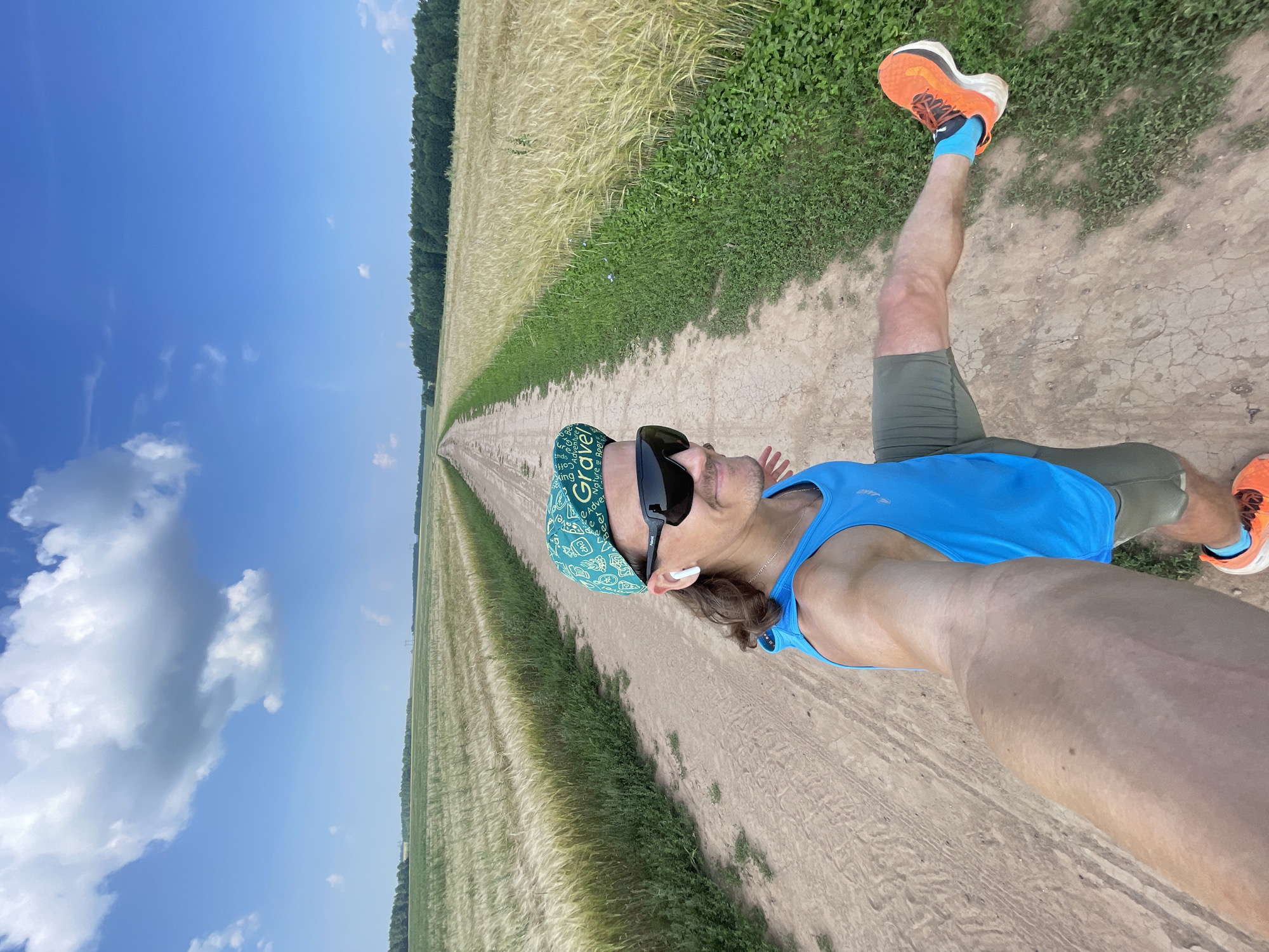 An individual wearing a blue tank top, olive green shorts, and orange running shoes is captured in a selfie on a dirt road in Maksimovo, Russia. The person is also wearing a green patterned cap and sunglasses, with earbuds in their ears. They are in mid-stride, with their right leg extended. The road is bordered by a field of dry, golden grass on the right and a similar field on the left, with a line of trees in the distance. The sky is a clear blue with scattered white clouds.
