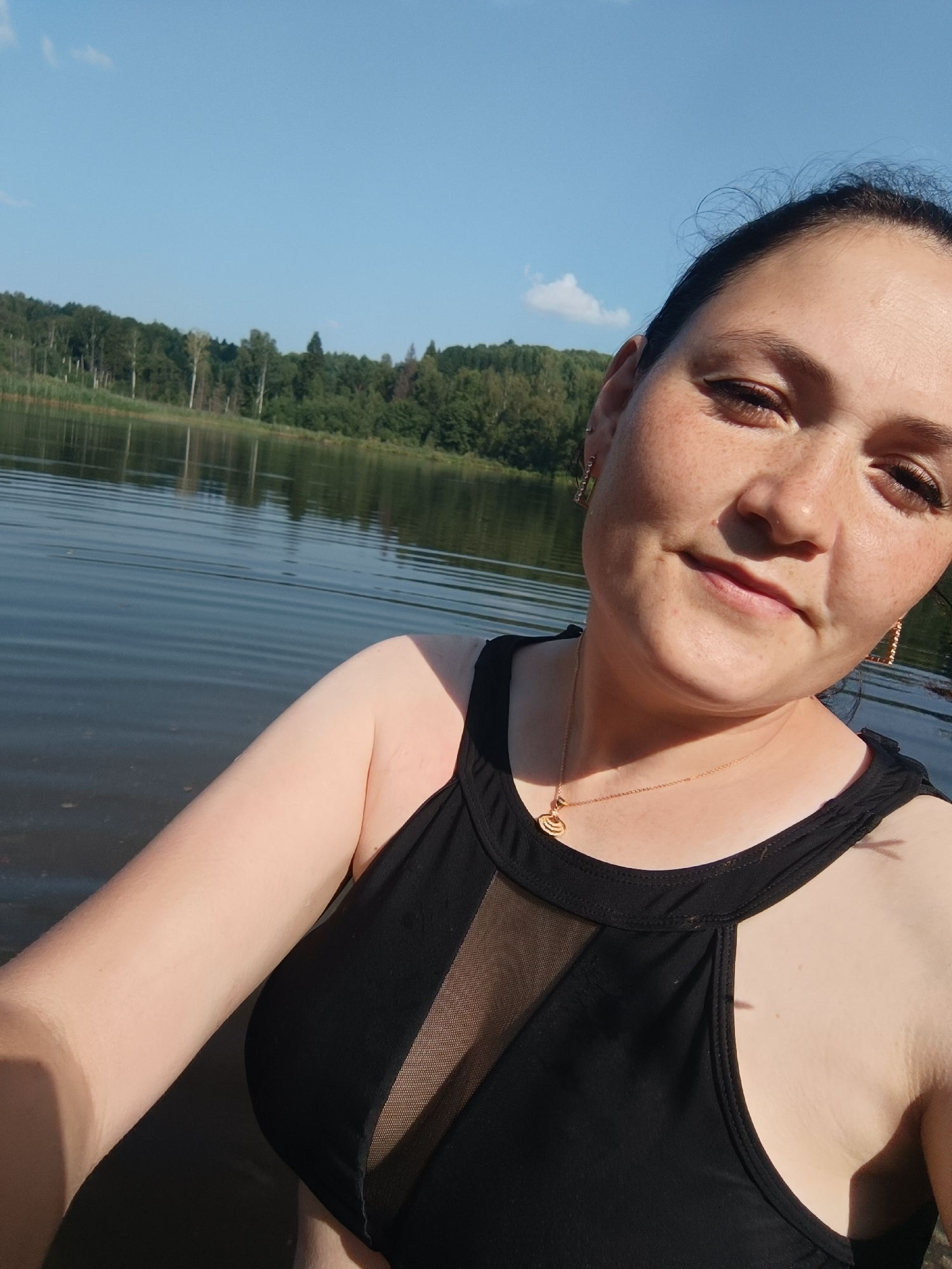 In a medium shot, a woman with dark hair pulled back and freckles across her nose and cheeks poses for a selfie on a body of water, likely a lake. She is wearing a black swimsuit with a mesh panel over the bust and a gold necklace with a circular pendant. The background shows a lush, tree-lined shore under a clear blue sky with a few wispy clouds. The water is calm with ripples reflecting the sunlight. The scene suggests a recreational setting, possibly a park or natural reserve. The specific location in the city of Karsha, Russia, is not explicitly identifiable from the visual cues provided in the image.