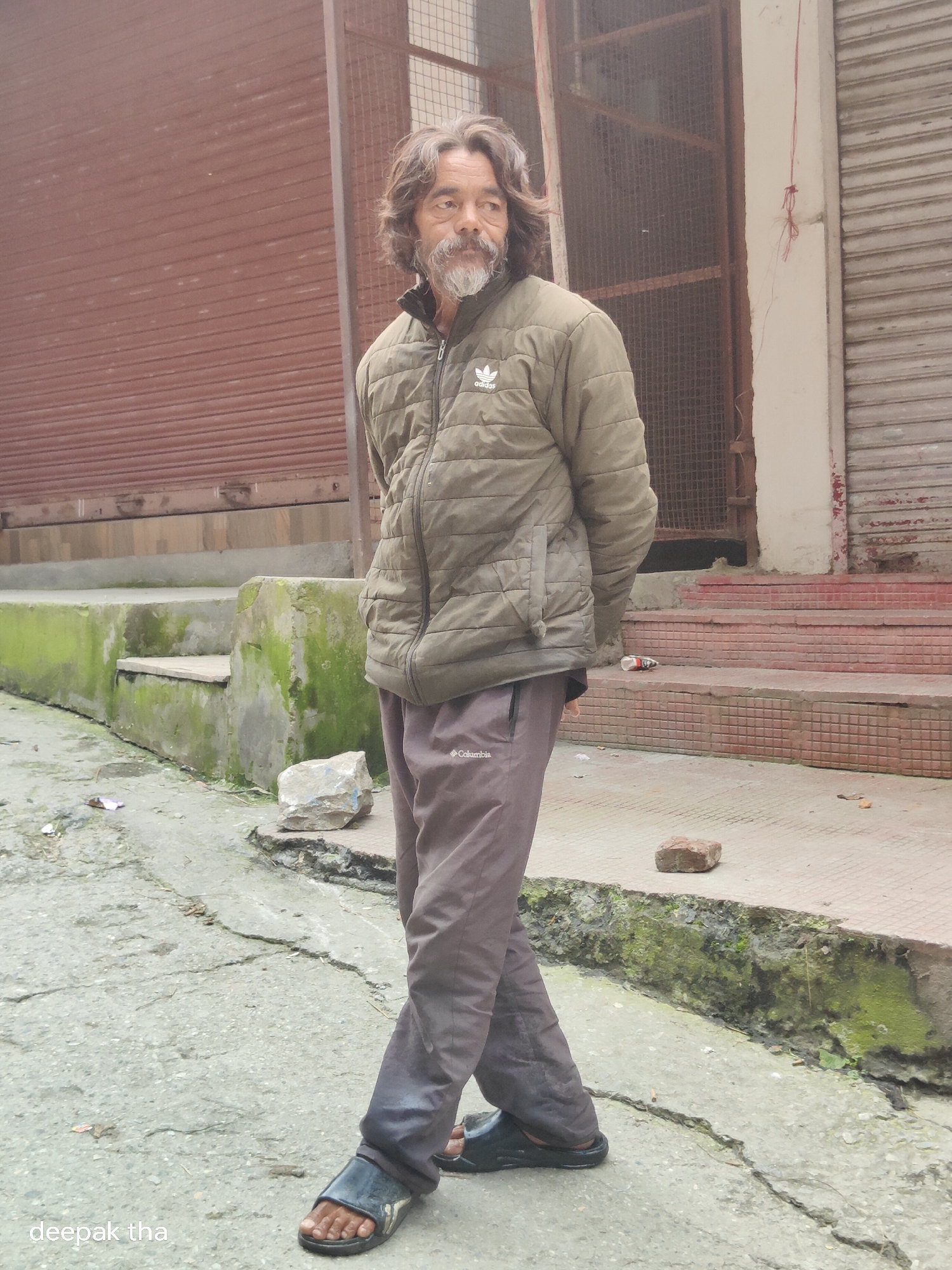 An elderly man with long, unkempt grey hair and a beard stands on a cracked, weathered concrete path in Vashisht, India. He is wearing a dark olive green puffer jacket with the Adidas logo on the left chest and dark grey athletic pants with the Columbia logo on the right thigh. His left leg is crossed over his right, and he is looking to his right. He is wearing black slip-on sandals.
Behind him, a set of concrete stairs is covered in green moss and grime. The stairs lead up to a building with closed metal shutters, one a dark reddish-brown and the other a lighter metallic color. The entrance area of the building has red tiled steps. In the foreground, scattered on the path and stairs, are rocks, debris, and some dried leaves. A white wrapper is visible on the path to the left of the stairs. The overall environment appears slightly damp and worn. The sky is overcast, creating diffused light. A watermark with the text "deepak tha" is visible in the bottom left corner.