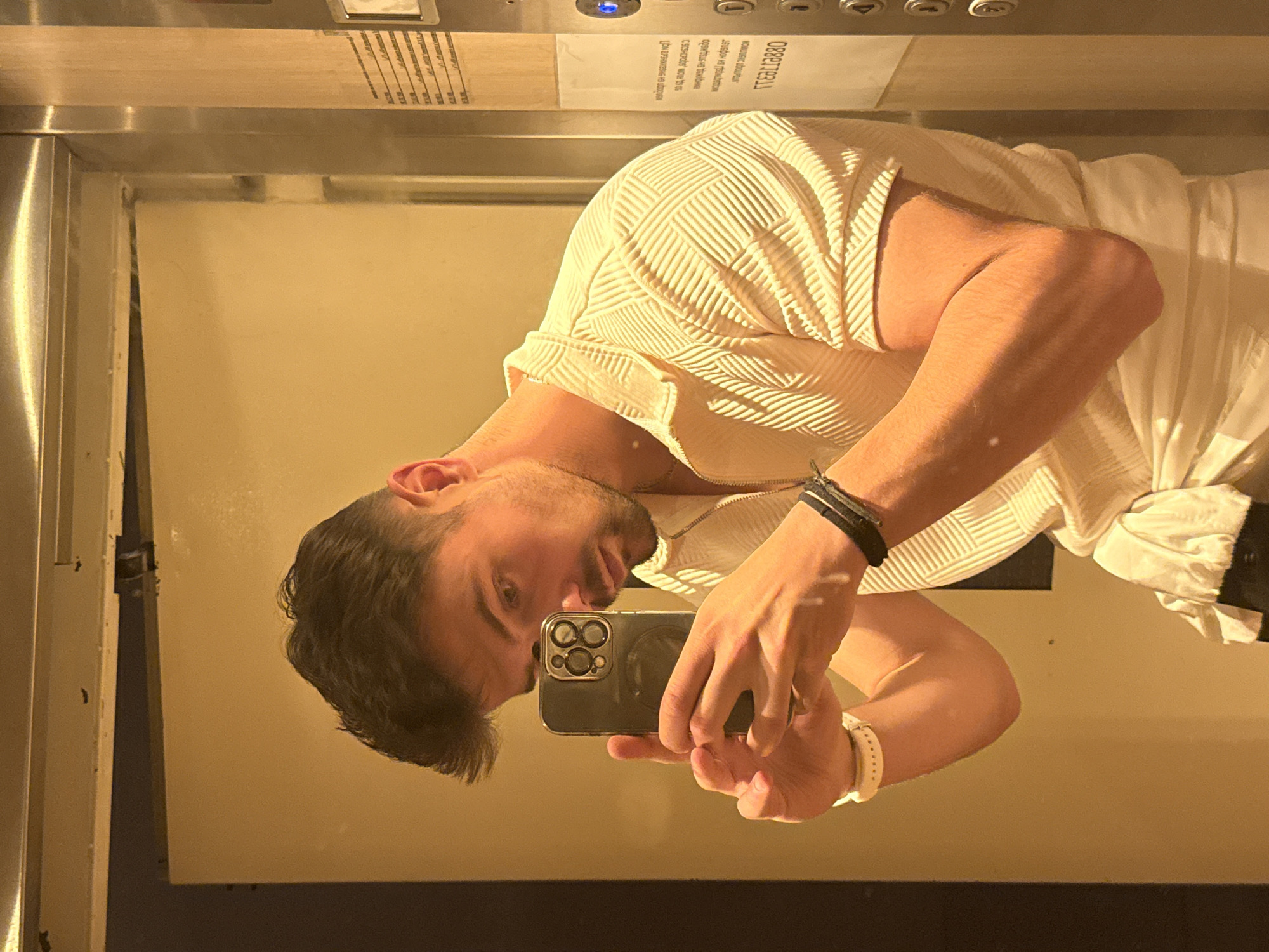 The image displays a rotated reflection of a man taking a selfie within what appears to be an elevator in Sofia, Bulgaria. The original image seems to have been taken as a vertical mirror selfie and then rotated 90 degrees clockwise.
The man, seen from the chest up in the reflection, has dark hair and a beard. He is wearing a light-colored, possibly cream or off-white, short-sleeved shirt featuring a distinctive textured, geometric pattern. On his left wrist (which appears on the right side of the reflection), he wears a black bracelet, and on his right wrist (appearing on the left), a white watch or band. He holds a dark-colored smartphone, equipped with a prominent triple-lens camera system, in his right hand, focusing on the screen to capture the reflection.
The setting appears to be an interior space, likely an elevator, given the metallic frame visible on the left side of the reflection and the control panel elements at the top. The background is a warm, light-beige or light-brown wall. The lighting is artificial and casts a warm glow, suggesting an indoor environment, possibly in the evening or at night.
Several lines of text are visible as reflections on a white sign, indicating a Bulgarian context:
- "ОБЩИТЕЛЛИ" (upside down and mirrored, meaning "Contractors")
- "КОМАНДЕН МОМЕНТ" (upside down and mirrored, meaning "Command Moment" or "Team Moment")
- "забележки на потребителя" (upside down and mirrored, meaning "user notes")
- A sequence of numbers and letters, "088ТЕJ11" (upside down and mirrored), possibly a phone number or identification code.
The man's expression is neutral as he concentrates on taking the photograph.