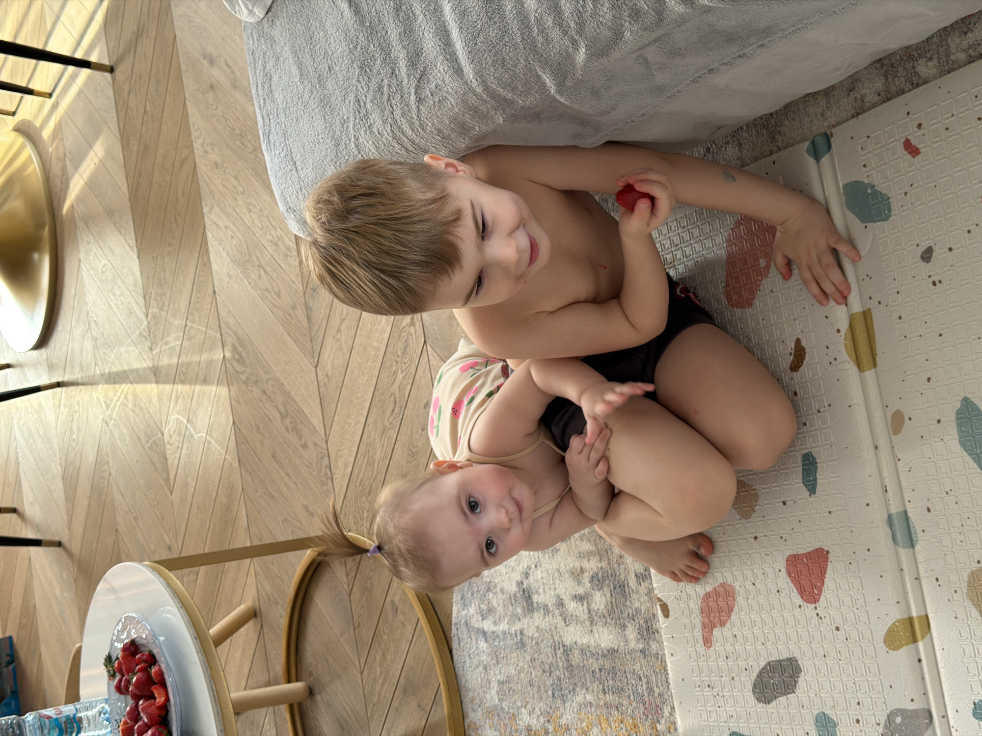 Two young children, a boy and a girl, are seated on the floor in a brightly lit indoor setting in Krasnoyarsk, Russia. The boy, with short blonde hair, is shirtless and wears dark bottoms. He holds a red strawberry in his left hand and looks down with a happy, smiling expression. Behind him, the younger girl, with light brown hair tied in a ponytail with a purple hair elastic, wears a light-colored top with a strawberry pattern and dark bottoms. She is looking directly at the camera with a gentle, pleasant expression.
The children are positioned on a transition of floor coverings: light-colored wooden herringbone parquet flooring is on the left, leading to a patterned rug beneath the girl, and then to a light-colored playmat with geometric textures and colorful abstract shapes (red, blue, yellow, grey) on the right. In the upper right background, a grey textured blanket or sheet from a bed or sofa is visible.
To the far left, a small round table with a golden base and white top holds a white bowl overflowing with fresh strawberries and a plastic water bottle. Other golden, metallic decorative elements are also visible on the left. Bright natural light streams into the room, casting distinct shadows on the wooden floor, suggesting it is daytime. Both children appear content and engaged in their activity, possibly snacking on strawberries. The text on the water bottle label is unreadable.