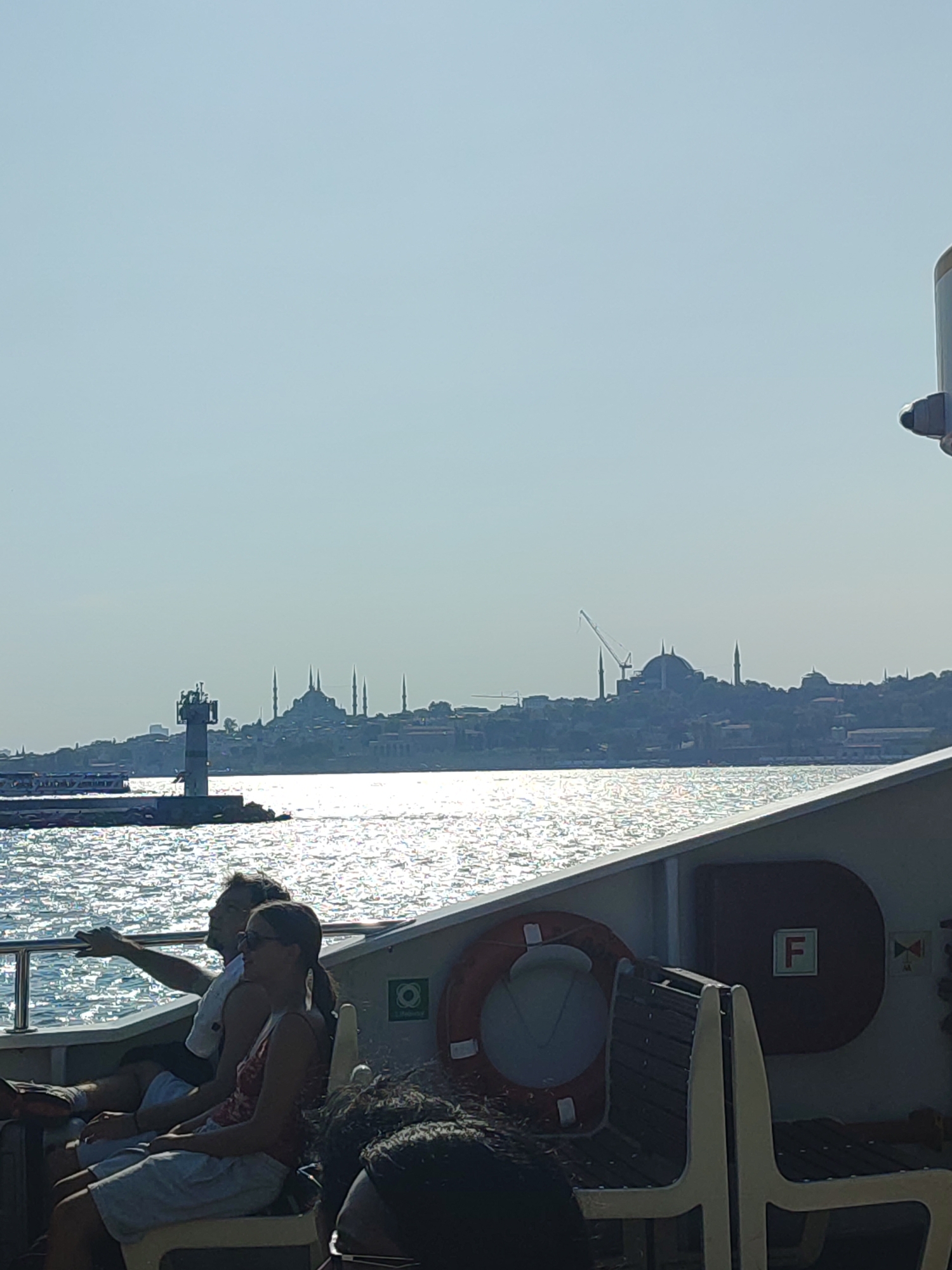 In Üsküdar, Turkey, a ferry carries passengers across the Bosphorus Strait under a clear sky. In the foreground, two people, a man and a woman, are seated on the deck of the ferry, looking out at the water. The man, wearing glasses and a white shirt, points towards the distant shore. The woman, with her hair in a ponytail, wears sunglasses and a patterned tank top. Behind them, a life preserver is attached to the side of the ferry, along with a red box with the letter "F" in white. The water is choppy and reflects the sunlight, creating a shimmering effect. In the background, the silhouette of the Istanbul skyline is visible, dominated by the domes and minarets of mosques, including the Hagia Sophia. A lighthouse stands on a rocky outcrop in the middle distance, and other ferries can be seen on the water.