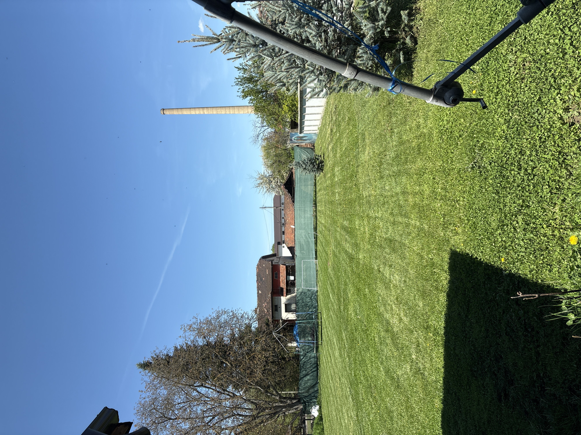 The image shows a grassy backyard in Kopilovtsi, Bulgaria on a sunny day. A tall, brick smokestack is visible in the distance. A fence separates the yard from adjacent houses. A trampoline and a small soccer goal are set up near the fence. A tree without leaves and a section of roof are visible in the lower left corner. A shadow is present in the lower right corner of the yard. There are lawn trimming marks on the grass. An object possibly related to lawn care with a blue rope is positioned in the upper portion of the frame.