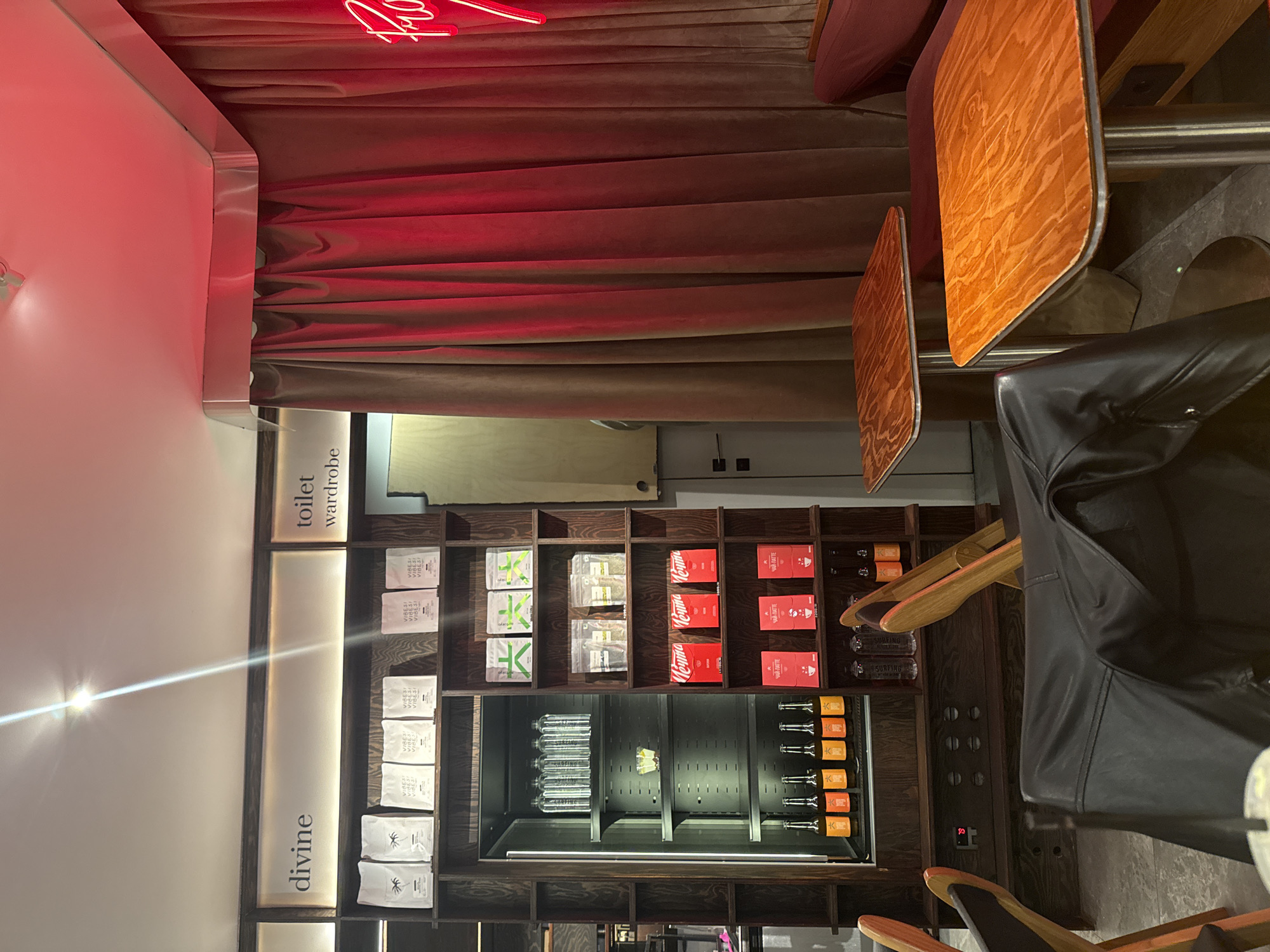 The image depicts the interior of a cafe or bar located in Yerevan, Armenia. The scene is dominated by a dark wooden shelving unit on the left, displaying various products. The top shelves feature white pouches labeled "VIBES!" with a palm tree logo, while lower shelves contain green-labeled pouches, clear pouches, red boxes marked "Keyma," and red/black boxes labeled "YAR-RATTE." An integrated, glass-fronted cooler section within the shelving holds multiple clear bottles on the upper shelf and several orange-labeled bottles on the bottom shelf. Above the shelving, backlit signs read "divine" and "toilet wardrobe."
To the right, a heavy, floor-to-ceiling brown curtain, illuminated by a red light source, likely a neon sign partially visible on the far right, covers a wall. Behind the curtain, a light-colored wooden door with a dark handle is visible, corresponding to the "toilet wardrobe" area. In the foreground and midground, two square tables with polished wooden tops and chrome pedestal bases are arranged. A dark red upholstered booth-style seat is visible behind the rightmost table. A black leather jacket is draped over the back of a dark chair in the immediate foreground, partially obscuring a blurry white cup with a straw. The floor appears to be a light-colored, possibly concrete or tiled, surface. No individuals are present in the frame.
