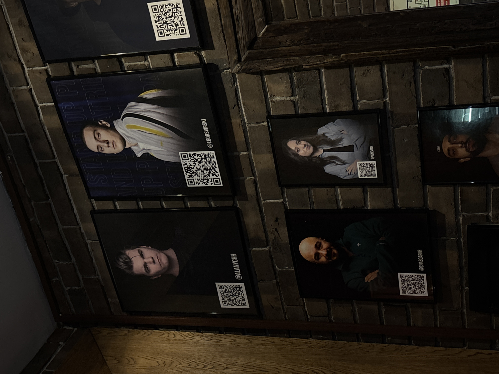 The image shows a brick wall in what appears to be an interior space, possibly in Moscow, Russia, based on the Cyrillic alphabet in the posters. Several framed posters are affixed to the wall. The posters depict headshots of various individuals, possibly comedians or performers, each with a QR code and an associated Instagram handle, such as "@EGORSVIRSKI," "@ALANYCHH," "@CHABDARRR," and "@VENEVITO". The posters are arranged somewhat haphazardly on the wall. The lighting is subdued, and the overall aesthetic is that of a promotional display in a bar or comedy club. Wood paneling is visible near the bottom left.
