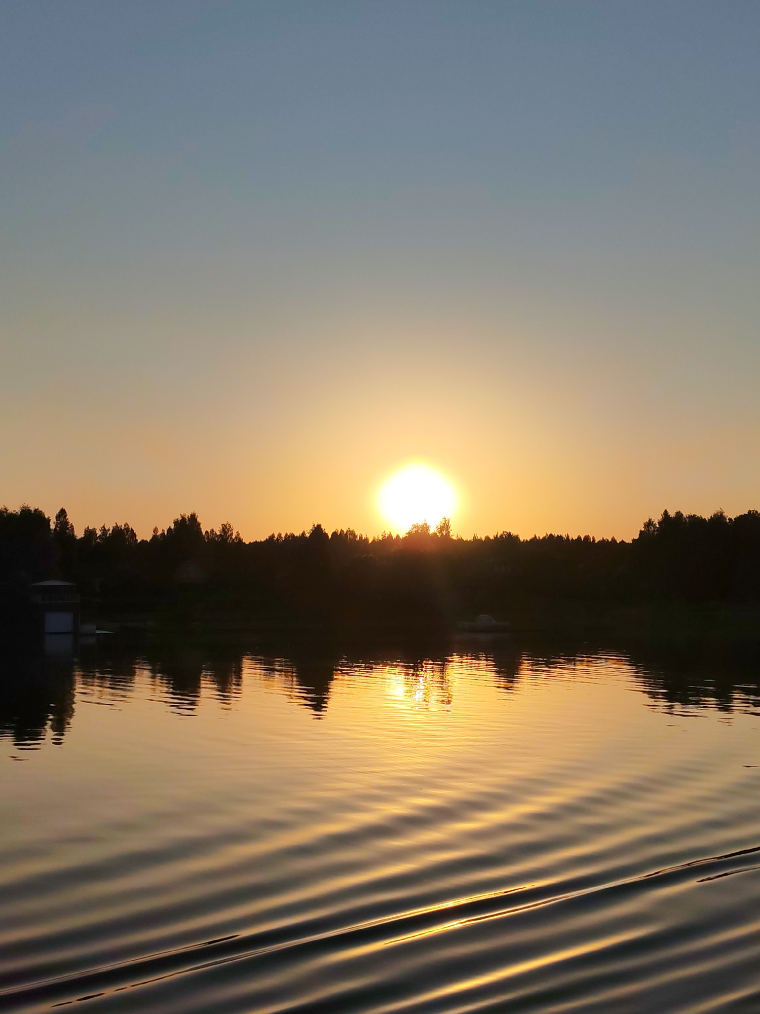 The image shows a sunset over a body of water, possibly a lake or river. The sun is positioned near the horizon, its light creating a golden reflection on the water's surface. Trees line the horizon, appearing as dark silhouettes against the bright sky. The sky fades from a light blue at the top to a warm orange near the horizon. Ripples or waves are visible on the water, adding texture and movement to the scene. The location and city are unknown.