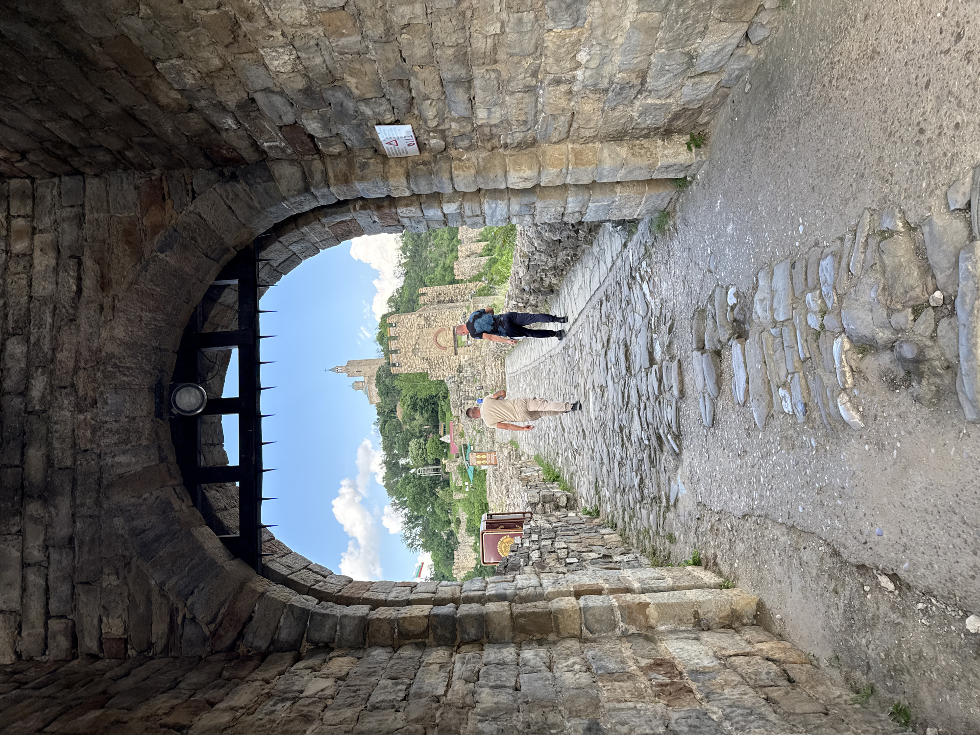 This photograph shows a view from within a stone archway leading to an exterior scene at Veliko Tarnovo, Bulgaria. Two people are present. One is walking away from the archway, wearing a tan outfit, while the other person, wearing black, is further away and appears to be taking a photo or looking at something. The walkway is paved with stones. The background includes parts of the city and a blue sky with some clouds. A metal portcullis partially obscures the view through the arch. There is a light fixture and a small sign on the stonework inside the arch.