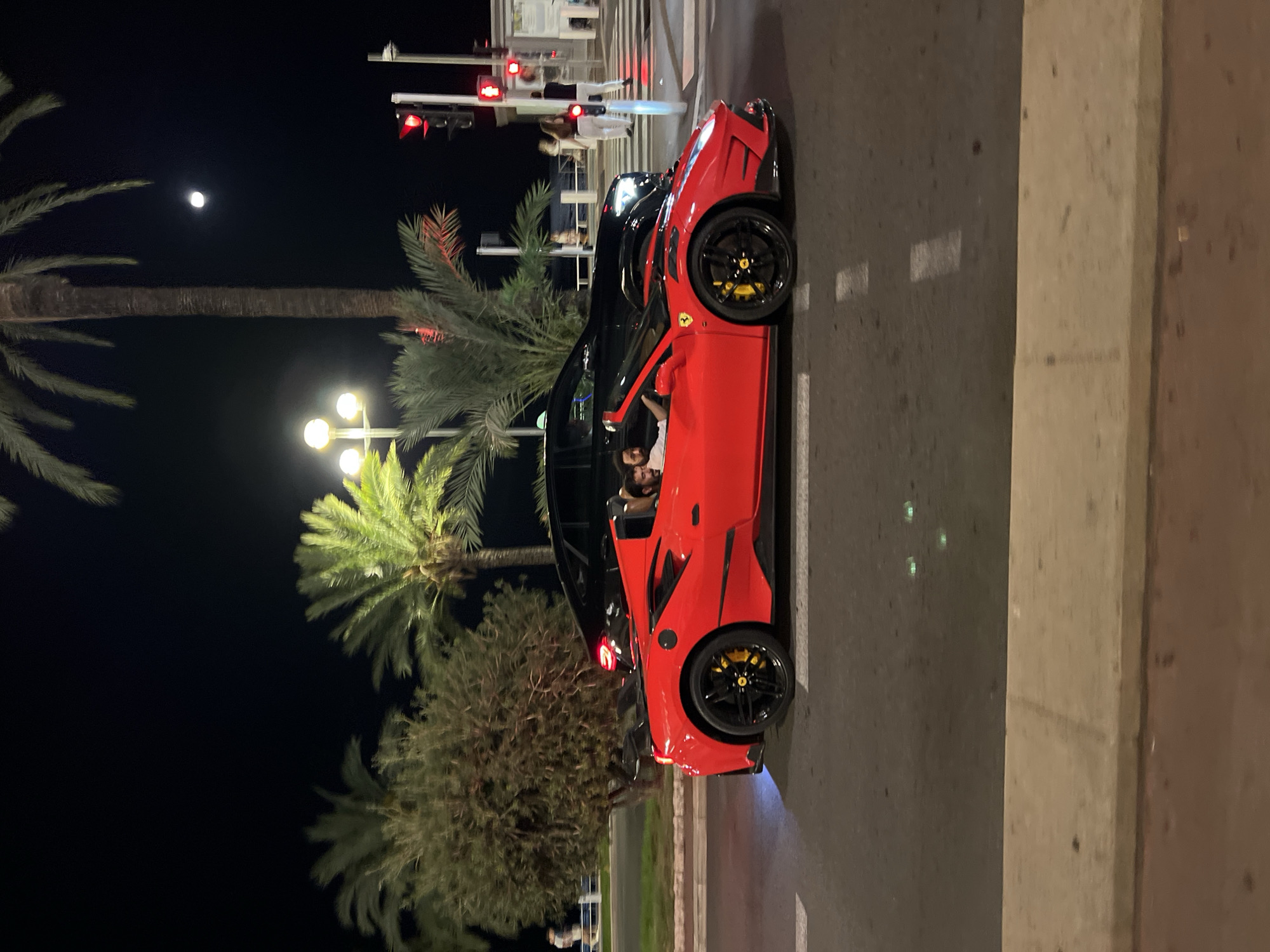 This is a nighttime, eye-level, medium shot of a bright red convertible sports car, likely a Ferrari, driving on a street in Nice, France. The car is in motion, with its headlights off, and its interior lights are on. Two men are visible inside the car; one is driving and the other is in the passenger seat. Both appear to be casually dressed.
Behind the red car, a dark-colored SUV or sedan is visible, also in motion. The road appears wet, reflecting streetlights and the car's taillights, suggesting recent rain or damp conditions. In the background, there are palm trees, streetlights, and traffic signals indicating a city setting. The traffic lights are red, suggesting the vehicles are approaching an intersection. Several people are visible on the sidewalk in the distance, some walking and some standing near a crosswalk. The overall atmosphere is that of a vibrant, illuminated urban environment at night. The presence of palm trees and the Mediterranean feel suggest this location is likely along the Promenade des Anglais in Nice.