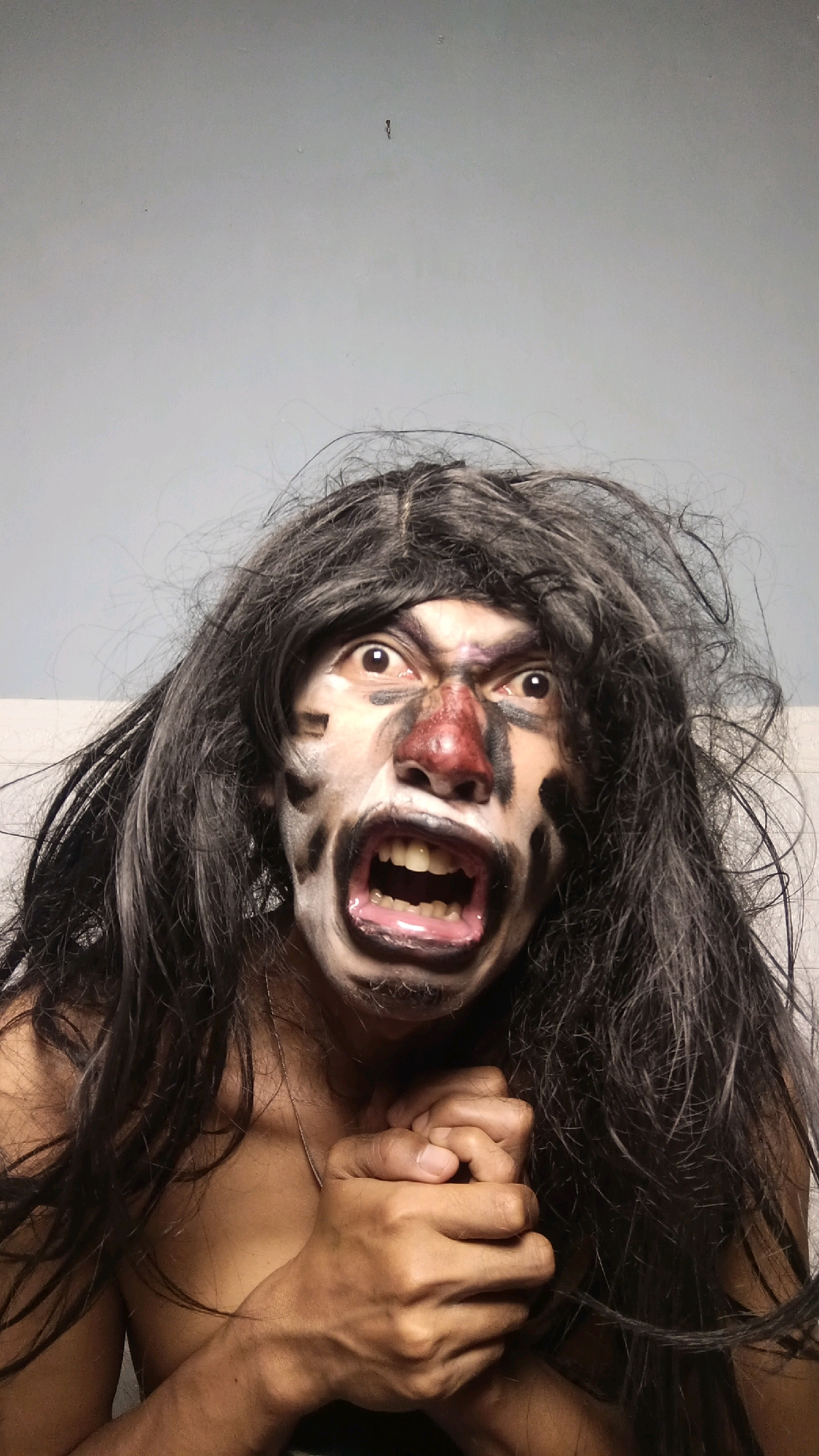 A person is posing for a photo with their face painted in a tribal or warrior-like style, with black and white markings and a red nose. They have long, dark, disheveled hair and are wearing makeup that exaggerates their features, including wide-open eyes and a screaming mouth showing teeth. Their hands are clasped together in front of their chest. The background is a plain, light-colored wall. The image appears to be a self-portrait, likely taken indoors. There is no indication of the location in Kebumen, Indonesia.