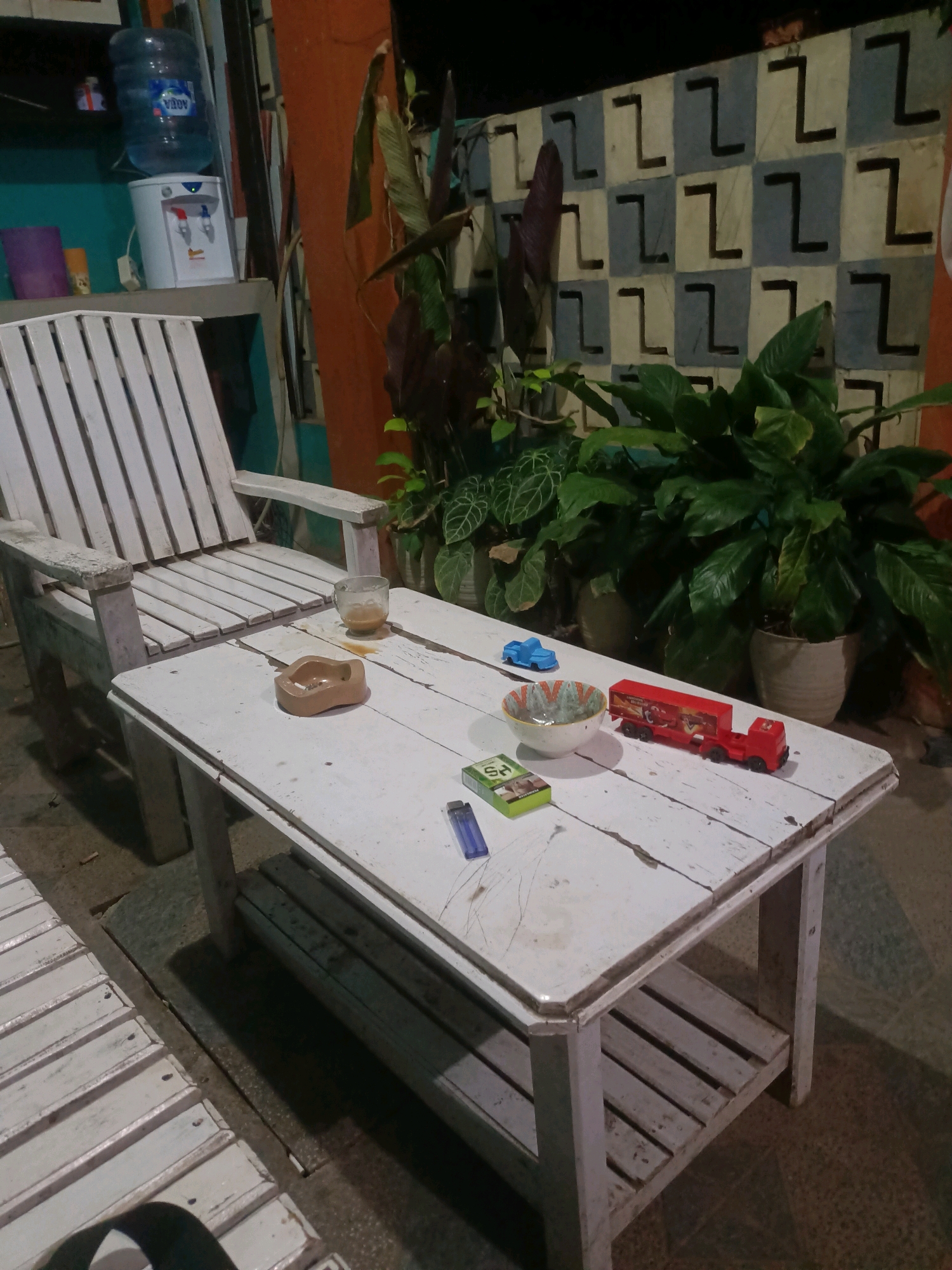 An outdoor seating area in Purwakarta, Indonesia, is depicted in the image. A weathered white wooden chair with slats is positioned to the left, and a matching wooden coffee table sits in front of it. On the table are several objects: a small glass with a brown liquid, an ashtray containing cigarette butts, a pack of cigarettes, a blue lighter, a blue toy car, a small bowl filled with liquid, and a red toy truck. Behind the table, a lush green plant is visible in a pot. The background features a patterned wall with alternating light and dark square tiles and a darker vertical orange element. The scene is illuminated by artificial light, suggesting it is nighttime.