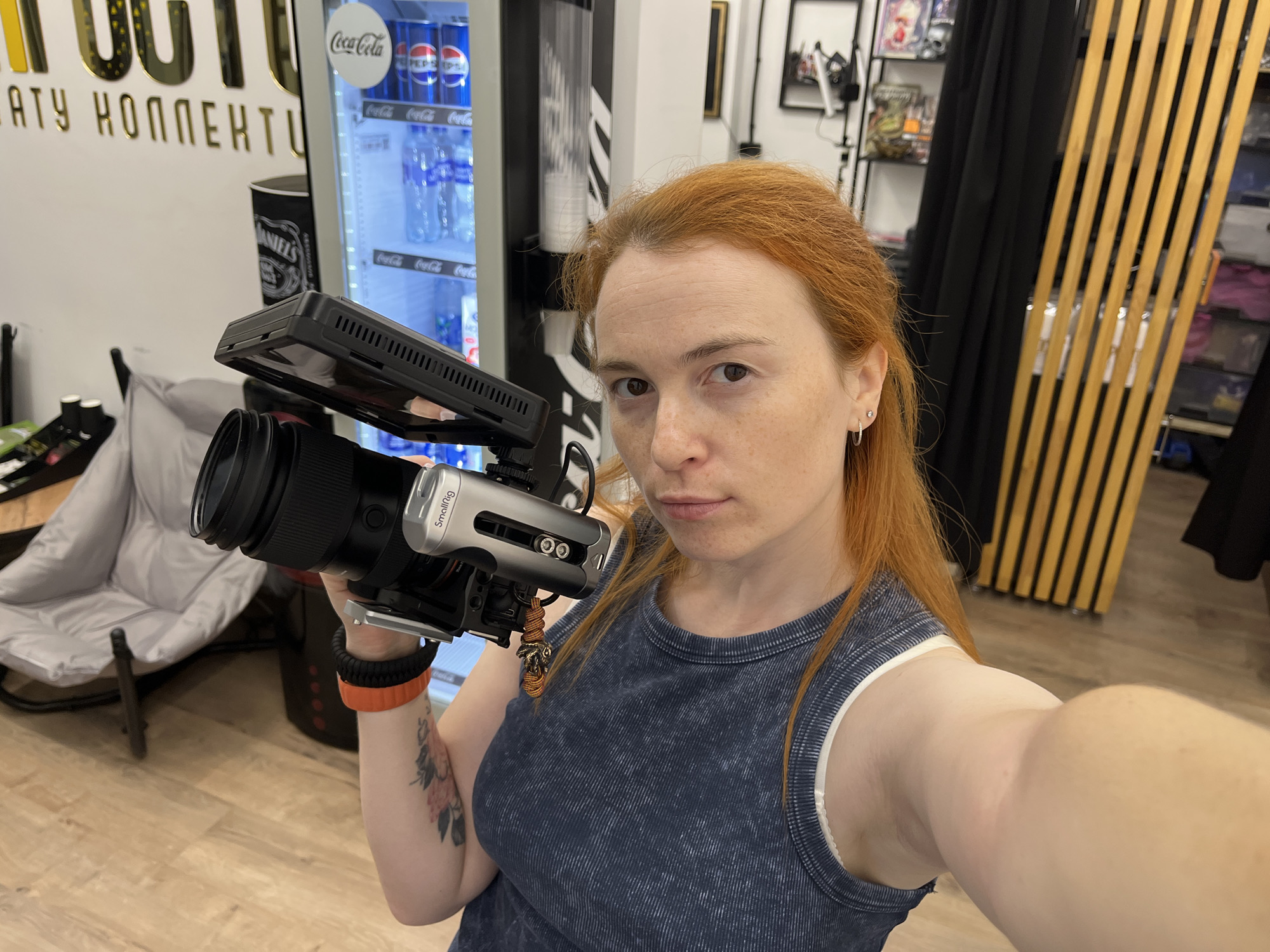 A woman with red hair is holding a camera with a large lens in her right hand. She is wearing a blue, ribbed tank top and a white bra. She has a tattoo of flowers on her left arm and is wearing a black and orange bracelet on her right wrist. Behind her is a cooler filled with Coca-Cola and Pepsi products, a black curtain, and wooden slats. The floor is wooden. A part of a gold text sign is visible on the left. The scene appears to be indoors, possibly in a shop or a similar commercial establishment. The location is not specified as Chaikovsky, Russia.