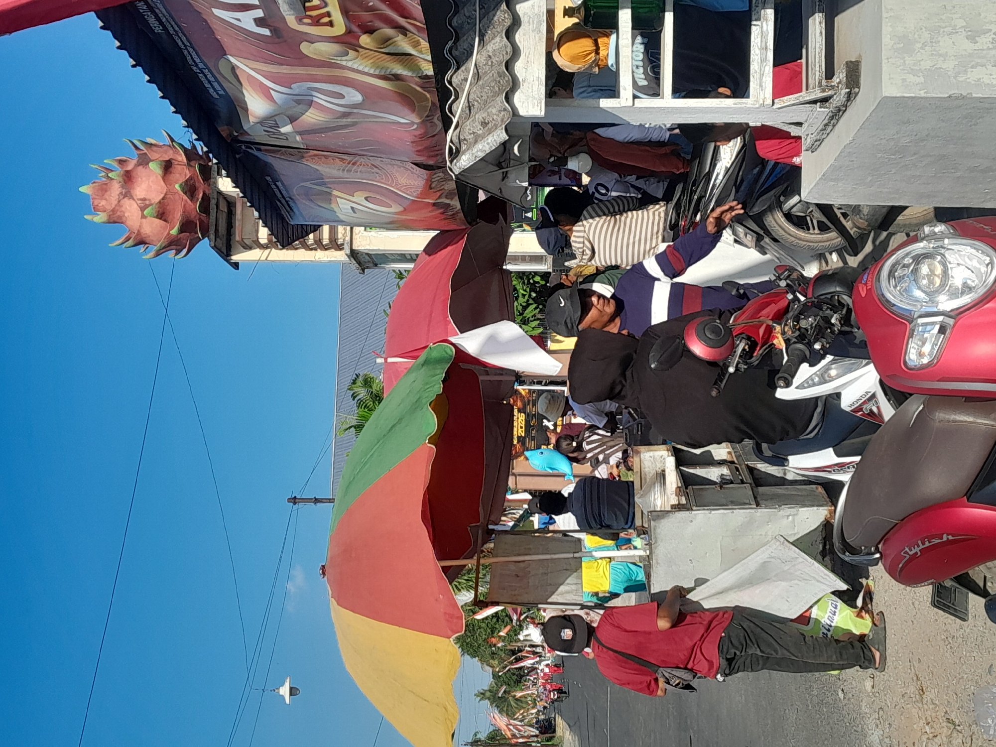 This is a street scene in Bulurejo, Indonesia, on a clear day with a bright blue sky. The image is dominated by several food stalls and a crowd of people.
In the foreground, a red scooter is parked, its front visible with a chrome headlight. To the left, a man wearing a red t-shirt and black pants stands next to a silver food cart, holding a white cloth. He is wearing a black fanny pack across his chest. Behind him, a large, colorful umbrella, divided into red, yellow, and green sections, shades the food stall. Another red umbrella, partially obscured, is also present.
Several people are gathered around the food stalls. A man in a black cap with a Nike logo is looking to his left. Another man wearing a striped sweater is reaching out his hand. A person in a black hooded jacket is seated on a scooter. Other individuals are visible in the background, some standing and some sitting.
Behind the stalls, a prominent building features a distinctive dragon fruit-shaped ornament on its roof, with green leafy tips and a reddish-pink body. A large banner with red and yellow graphics, possibly advertising food, hangs on the side of the building.
The street extends into the background, lined with trees and decorated with Indonesian flags. Several motorcycles and people are seen on the street, suggesting a bustling atmosphere. The ground appears to be a dirt or gravel surface. A street lamp hangs from a wire in the upper left corner of the image.