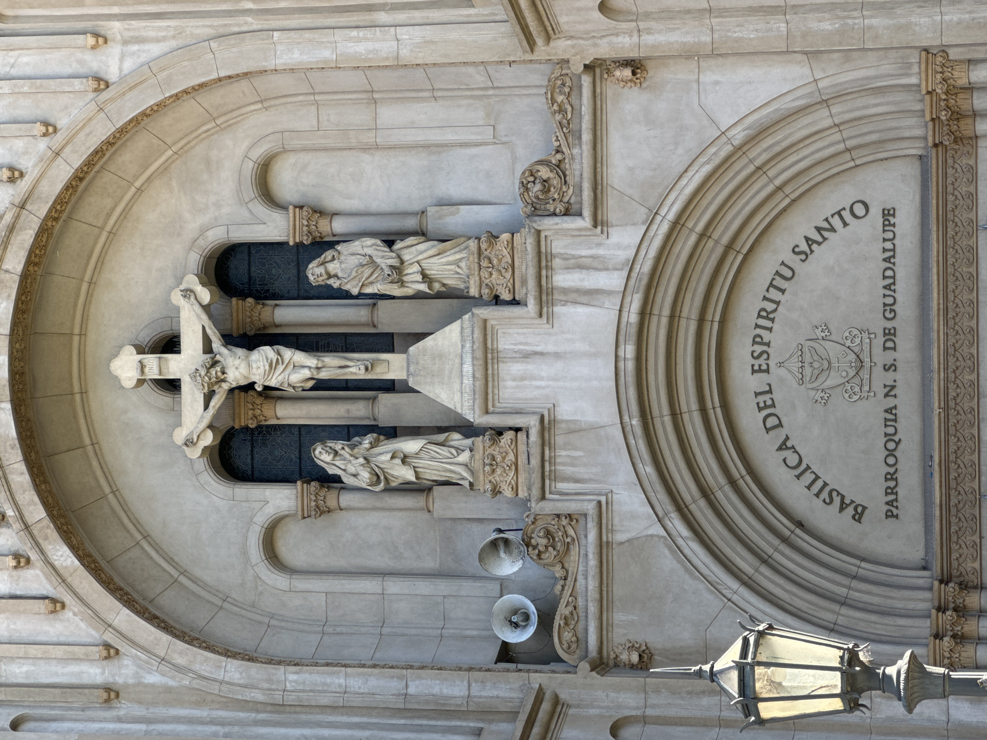The image shows a detailed architectural view of the Basilica Del Espiritu Santo Parroquia N. S. de Guadalupe in Buenos Aires, Argentina. The view is of a section of the facade featuring an arched niche containing a crucifix with a statue of a crucified Jesus figure. Flanking the crucifix are two additional statues. The architecture is light-colored stone or concrete with ornate carvings and moldings. A portion of the building's name and parish is inscribed on an adjacent arched surface. Below, a decorative lamp post is visible. Two speakers are also attached to the wall.