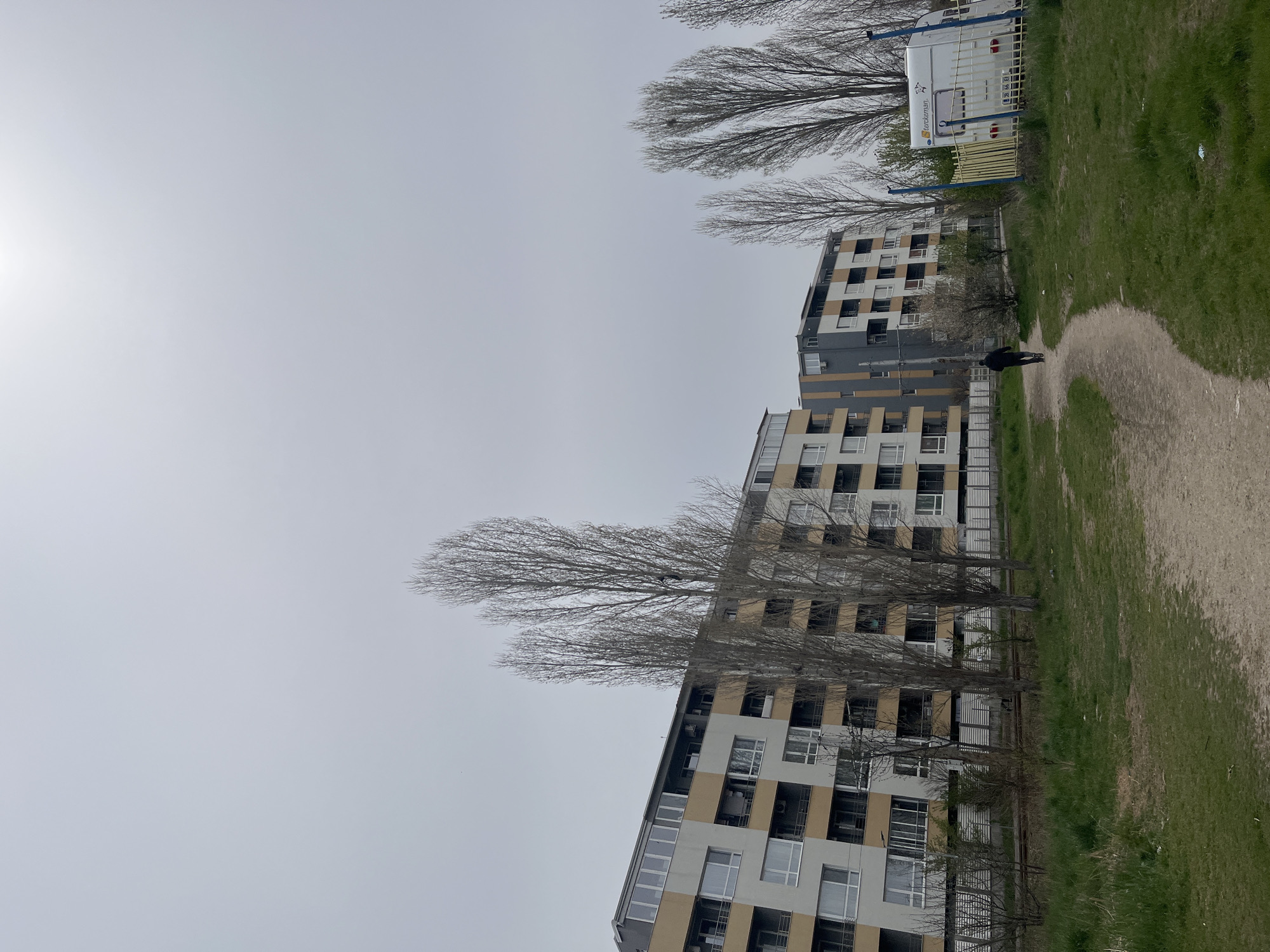 The image depicts a row of multi-story residential buildings in Sofia, Bulgaria. The buildings are characterized by a modern design with light-colored facades and dark window frames. Bare trees line the foreground, partially obscuring the view of the buildings. A gravel path runs parallel to the buildings, and a person is walking along the path. A white trailer or camper is parked behind the buildings, near a green grassy area. The sky is overcast and bright.