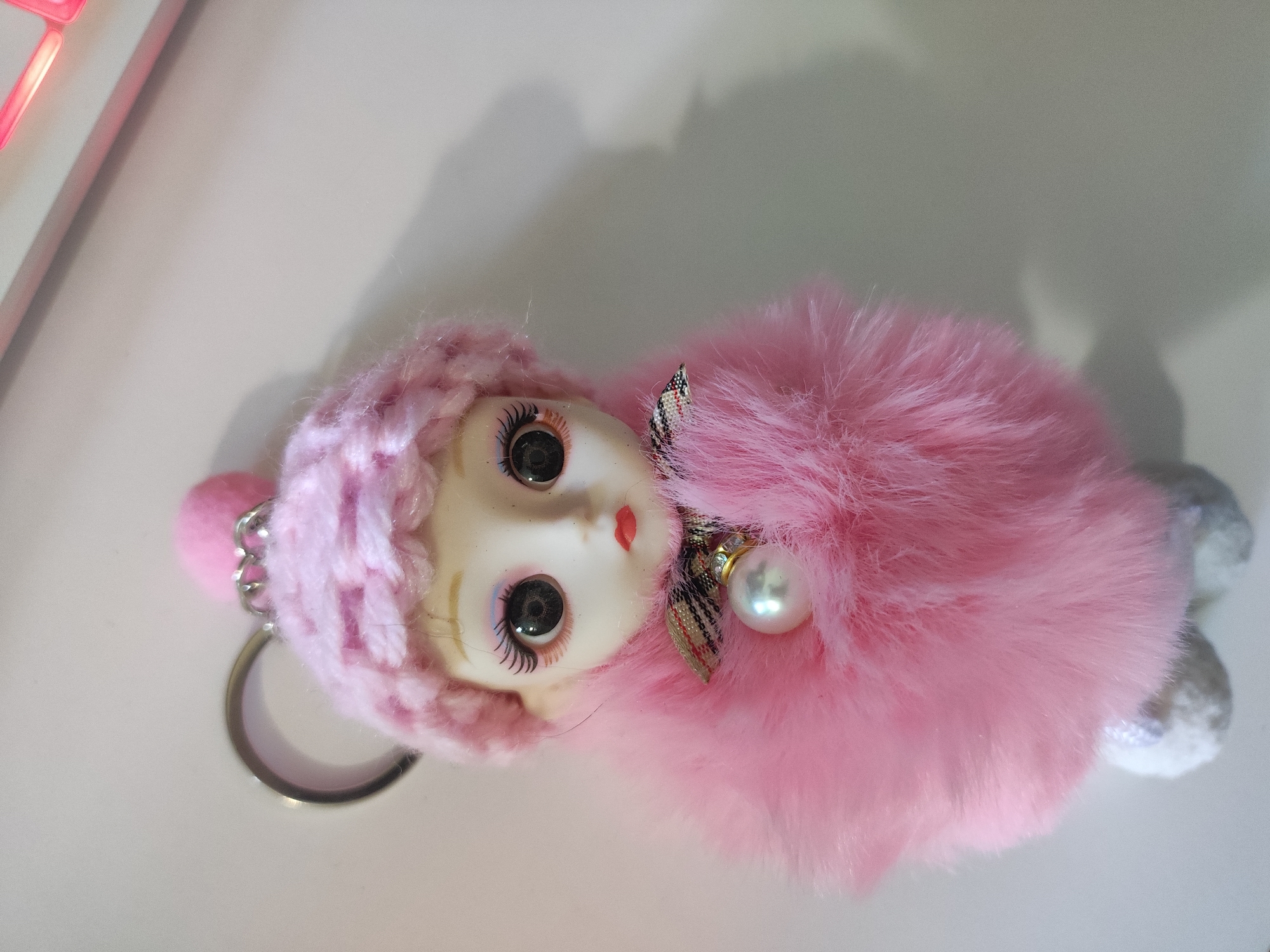 This is a close-up, eye-level, overhead shot of a pink fluffy doll keychain with a pearl accent. The doll has a pale face with wide, dark eyes, painted eyelashes, and red lips. It is wearing a pink knitted hat. The doll is attached to a silver keychain ring. The background is a plain, light-colored surface, likely a table or desk, with a shadow cast from the keychain. A blurred pink light source is visible in the top left corner. The image does not provide any information to suggest it is located in Cherkasy, Ukraine.