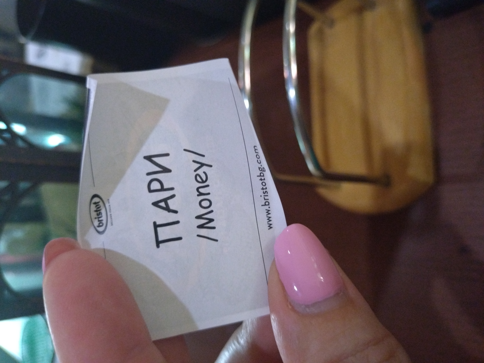 A close-up, slightly blurry shot shows a person's hand holding a white card with black text. The card is angled, with the top left corner obscured by a thumb and index finger. The background is a mix of dark and light areas, with a blurred yellow object and what appears to be a metal stand. The person's fingernails are painted a light pink.
The visible text on the card is in both Cyrillic and Latin alphabets. In Cyrillic, it reads "ПАРИ". Below this, in Latin script, it says "/Money/". There is also a logo with the word "brisfot" inside an oval, and a website address "www.bristotbg.com" further down the card. The card appears to be a tag or label of some kind, possibly relating to a product or service. The lighting is dim, suggesting it might be indoors during the day or evening. The overall impression is casual, with the focus on the card and the hand holding it.