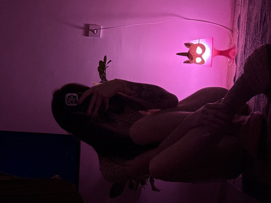 An individual poses for a selfie in a dimly lit room illuminated by pink light. The person holds a smartphone, obscuring their face, and has tattooed arms. A leopard-print garment is visible. The room features a unicorn-shaped lamp, a potted plant, a wall outlet, and what seems to be a television screen. The photo is rotated horizontally.