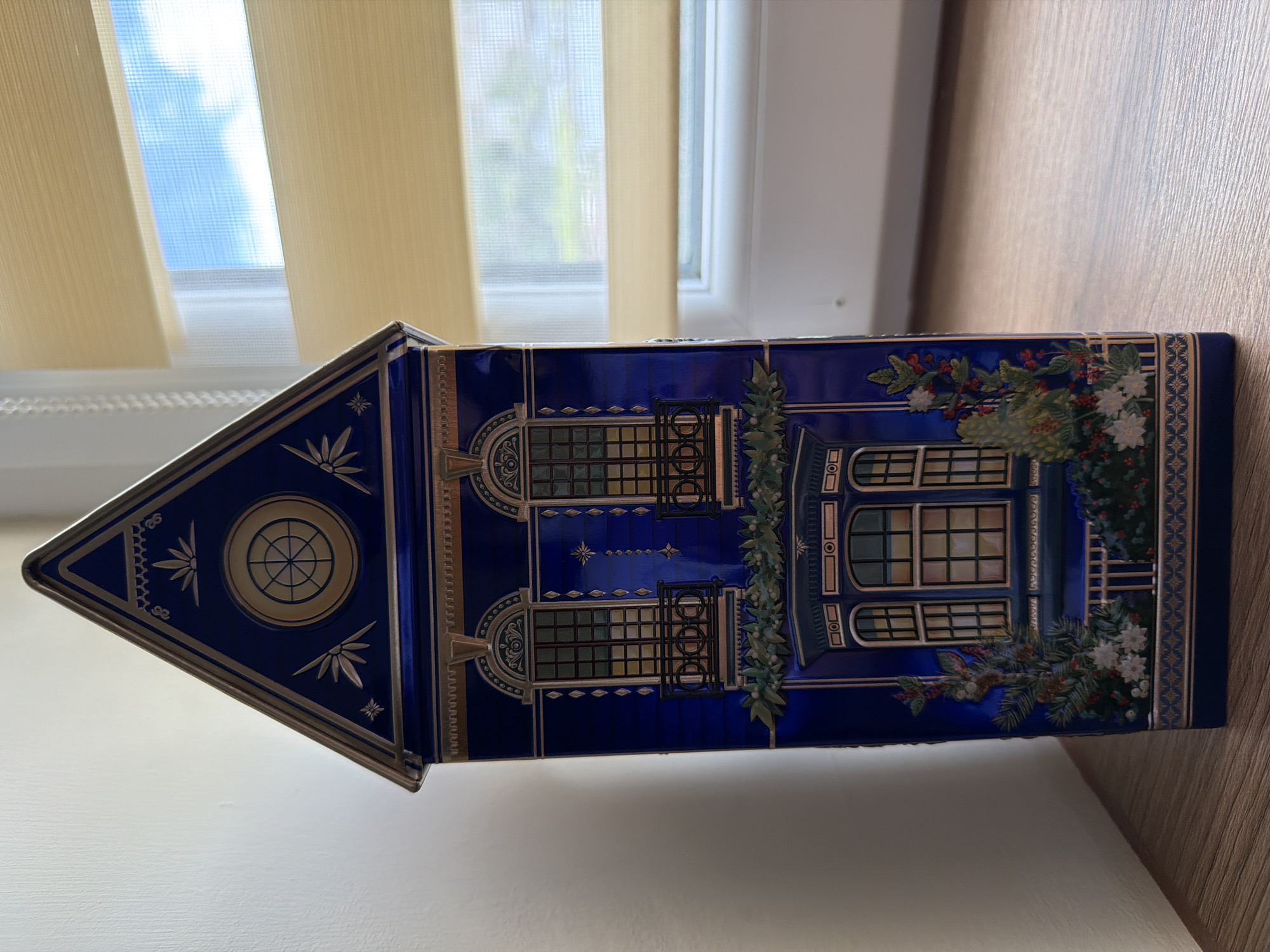A tall, dark blue, house-shaped tin is positioned on a wooden surface. The tin is decorated with gold trim and depicts architectural elements of a house, including windows with balconies, a wreath, and floral accents. A pale wall is visible to the left of the tin. To the right, a window with horizontal blinds is partially visible, showing a bright blue sky with clouds. The scene appears to be indoors, likely on a shelf or table, with the wooden surface extending to the right. There is no visible indication of the city of Pazardzhik, Bulgaria.