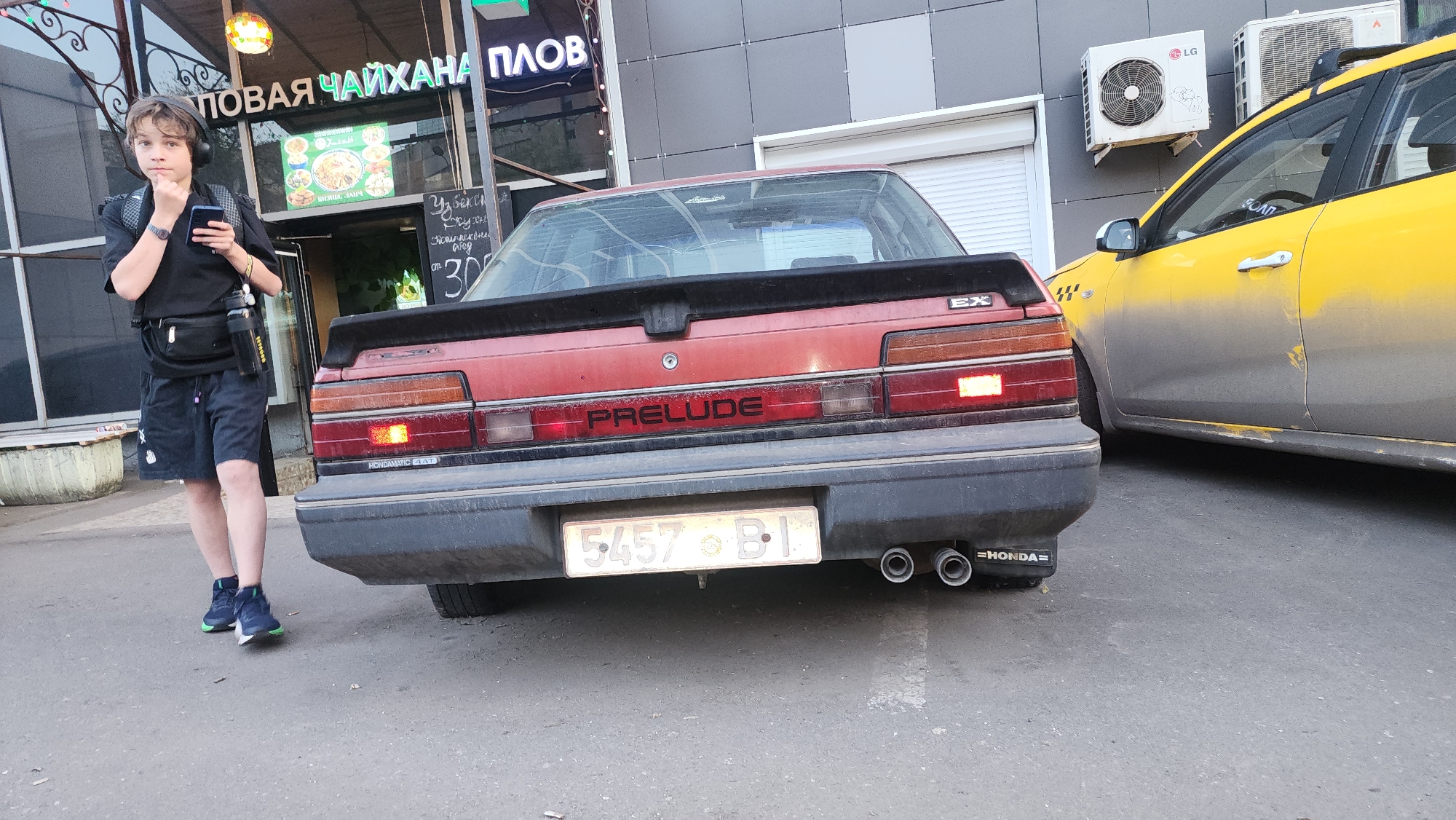 The image shows the rear of a red Honda Prelude EX parked on a city street. A young boy wearing headphones, a black t-shirt, dark shorts, and blue sneakers stands to the left of the car, looking at his phone. To the right of the Prelude is a dirty yellow taxi. In the background, there is a building with a sign that says "НОВАЯ ЧАЙХАНА ПЛОВ," which appears to be a restaurant specializing in plov. There is also a sign with Uzbek cuisine. Air conditioning units are mounted on the building behind the taxi. The license plate of the Prelude reads "5457 BI." The scene appears to be in Moscow, Russia.