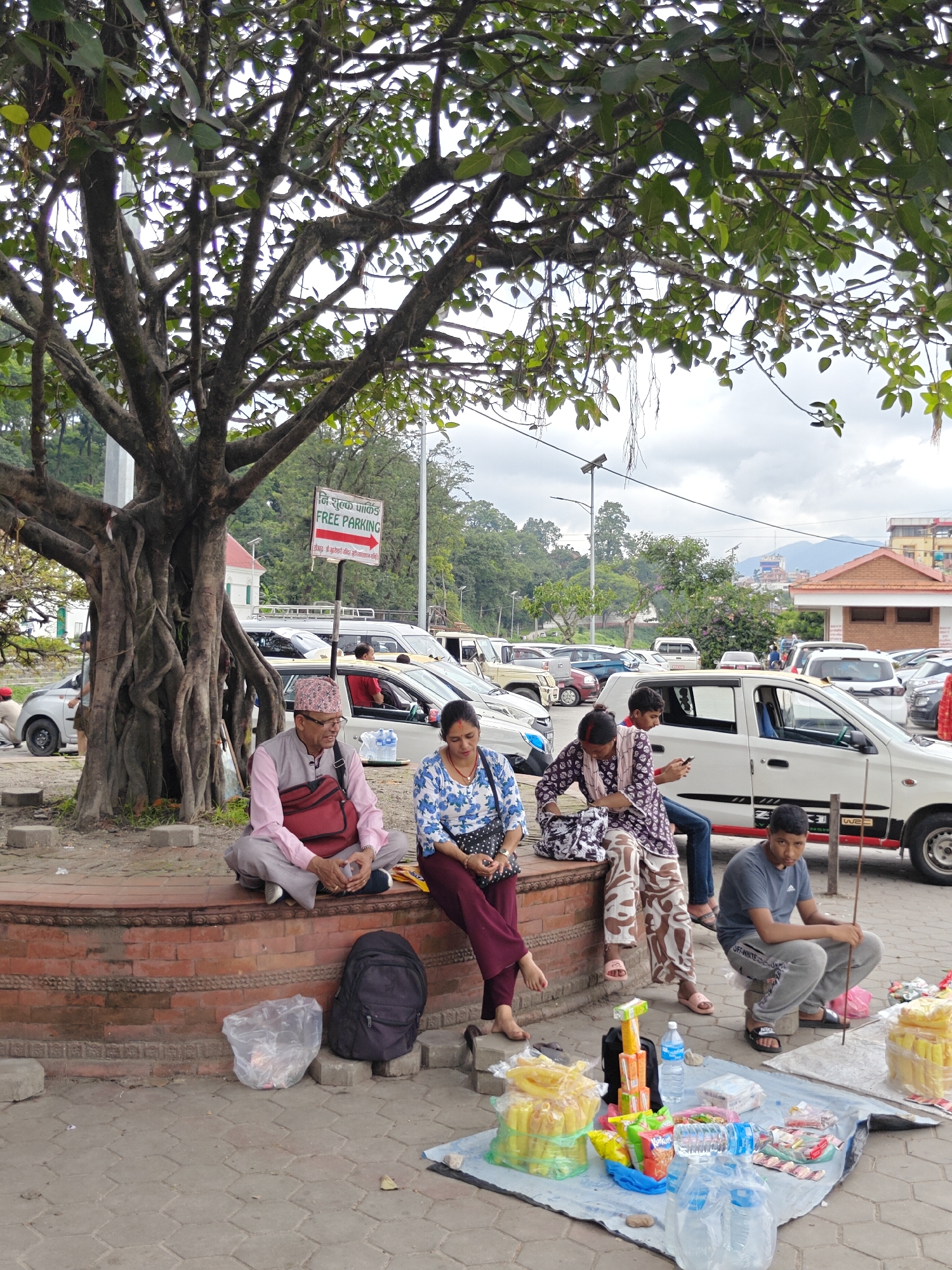 In Kathmandu Metropolitan City, Nepal, a street scene unfolds under the dappled shade of a large, gnarled tree. Several people are present in the foreground and midground. In the foreground, two individuals are seated on a low brick wall, with a third person to their right also seated. To the right of these individuals, a young man is crouched down near a stall set up on a blue tarp, displaying various packaged goods, including bottles of water and snacks. The stall appears to be a small vendor.
Behind the seated individuals, a white car is parked, and other vehicles are visible further in the background, suggesting a parking area or a street. A sign with Nepali script and an arrow pointing to the right is mounted on a pole, indicating "FREE PARKING." The environment is urban, with buildings and trees visible in the background under an overcast sky. The ground is paved with interlocking bricks. The overall atmosphere suggests a public space, possibly a roadside area or a market.