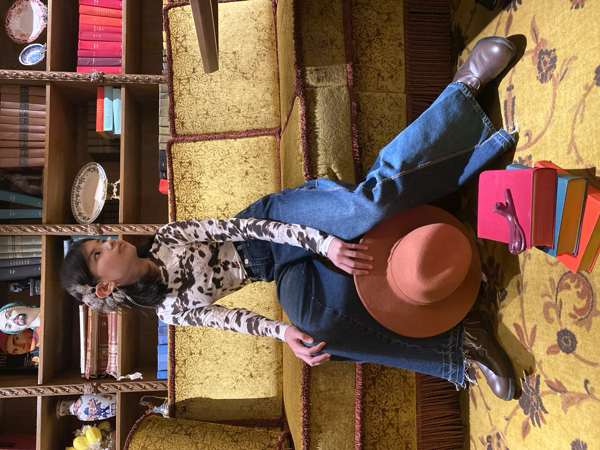 A young person is depicted lying on their back on a plush, golden-yellow sofa with maroon fringed edges, in what appears to be a richly decorated room, possibly in Yekaterinburg, Russia. The image is oriented such that the person appears to be lying vertically against the couch, which stands upright.
The subject, with dark hair, is looking upwards towards a wooden bookshelf that occupies the left side of the frame. They are wearing a long-sleeved shirt with a distinctive black and white cow print, blue wide-leg jeans, and dark brown boots. A rust-colored, wide-brimmed hat is held on their lap. A unique ear adornment, possibly made of feathers or fabric, is visible on their right ear. Their expression is calm and contemplative.
The sofa and the carpet beneath it both feature elaborate, repeating golden-yellow patterns, with darker floral or abstract motifs on the carpet. To the person's feet, a stack of vibrant books in pink, blue, and orange hues rests on the carpet, topped with a small, abstract purple sculpture.
The dark wooden bookshelf is meticulously filled with numerous books of varying sizes and colors, predominantly red, brown, and blue spines. Several decorative items, including ceramic plates with patterns, vases, and small statuettes (one resembling a bust with a painted face, another with lemons), are interspersed among the books. The overall setting suggests a cozy, intellectual, and perhaps antique-filled interior. No legible text is visible on the books or other objects.