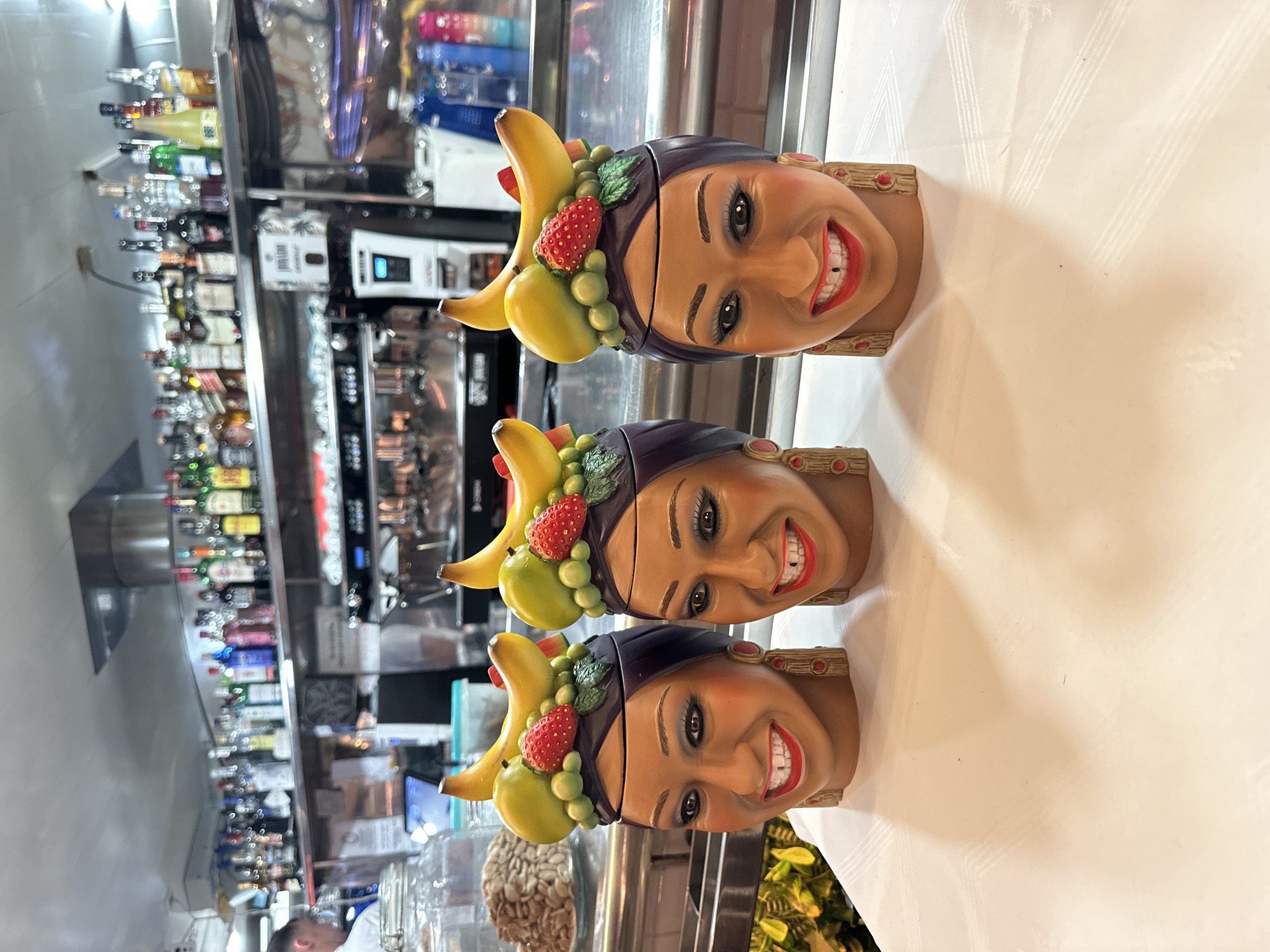 Three decorative ceramic head-shaped containers are displayed on a white tablecloth in the foreground. Each container depicts a woman's face with dark hair, large eyes, red lips, and a wide smile. The heads are adorned with artificial fruits, including bananas, strawberries, grapes, and a yellow fruit resembling a mango or pear. The containers are positioned in a row, close together. In the background, a bar is visible, stocked with numerous bottles of alcohol on shelves. A coffee machine and other bar equipment are also present. A person wearing a white shirt is partially visible on the far left. The overall setting appears to be a bar or restaurant.