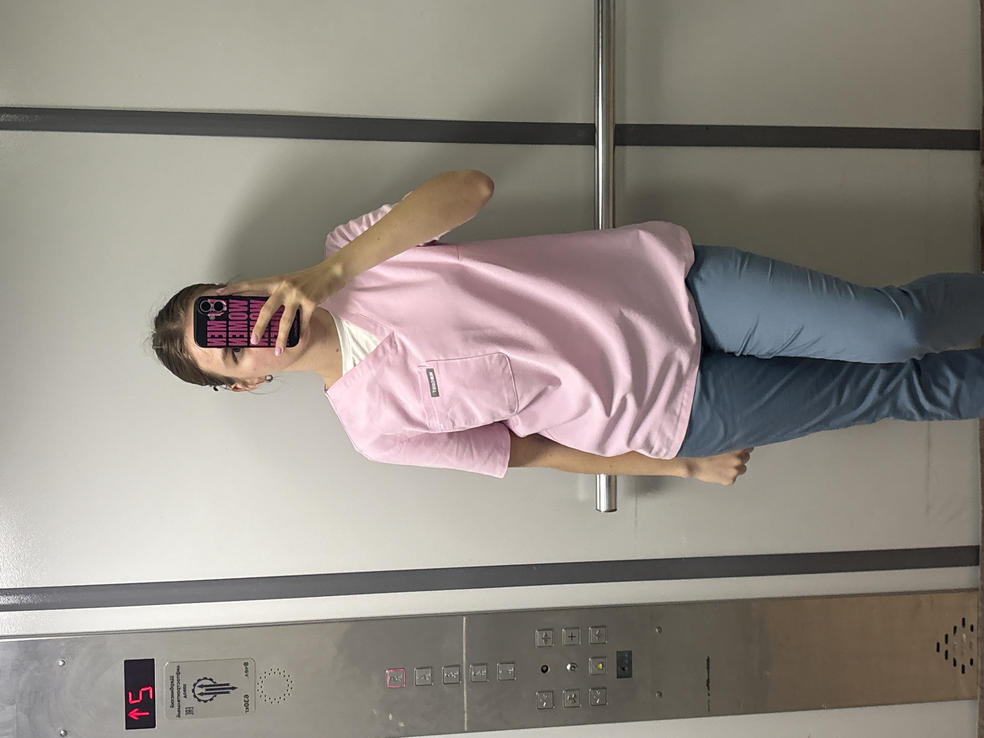 A young person in a pink scrub top and blue scrub pants is standing in an elevator, taking a selfie with a cell phone that obscures their face. The elevator's control panel is visible, indicating it is on the 15th floor. Text on the panel includes "Пермь" (Perm), which confirms the location as Perm, Russia. The elevator interior is plain with gray walls and a horizontal grab bar.