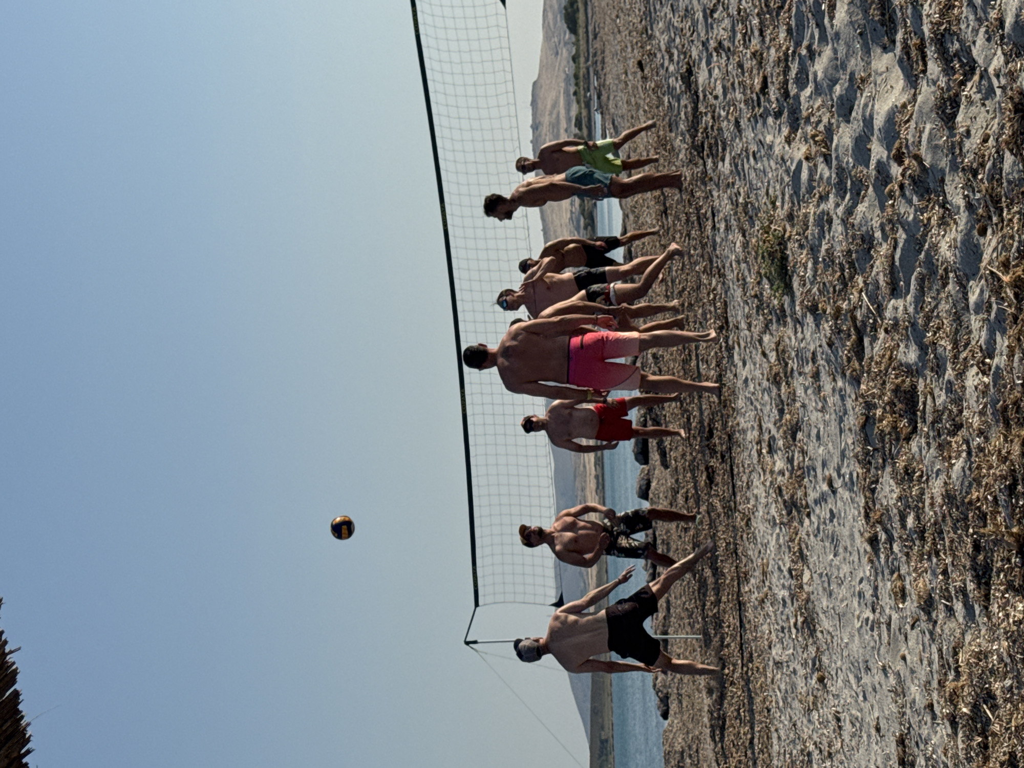 A group of men are playing beach volleyball on a rocky shore. There are at least eight men in the image, most of whom are shirtless and wearing swim trunks. The volleyball net is set up on the beach, and a volleyball is in the air, indicating an active game. The men are positioned on both sides of the net, with some in the process of hitting or receiving the ball. The ground is covered in pebbles and dry vegetation, typical of a Mediterranean coastline. In the background, the sea is visible, and beyond that, a hilly landscape. The sky is clear and blue, suggesting a sunny day, likely in the late morning or early afternoon. The overall mood appears to be one of friendly competition and recreation. The scene is set in Moudros Municipal Unit, Greece. There is no visible text in the image.
