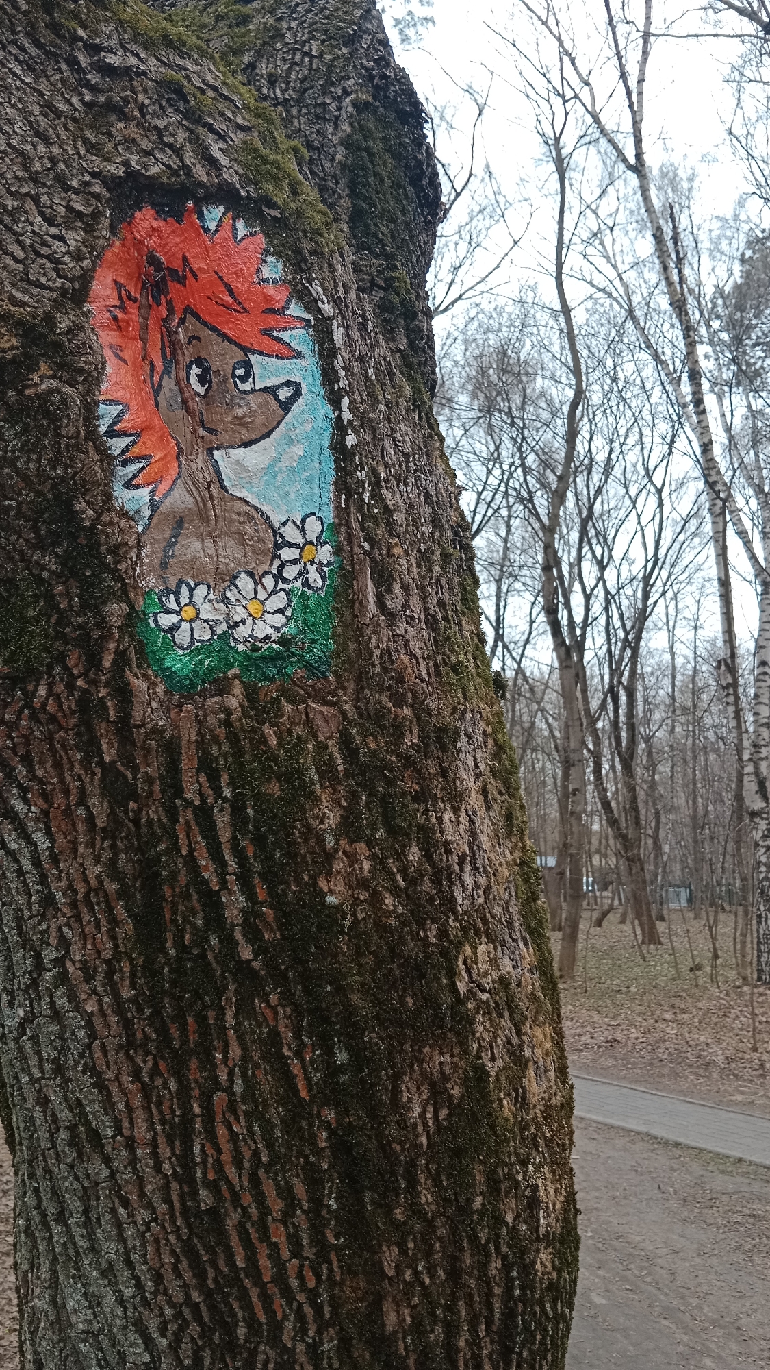 A painting of a hedgehog with red spikes on its head is on a tree trunk covered in moss in a park in Moscow, Russia. The hedgehog has big eyes and wears a collar of white daisies. The background of the painting is a light blue color. The park has bare trees and a paved path.
