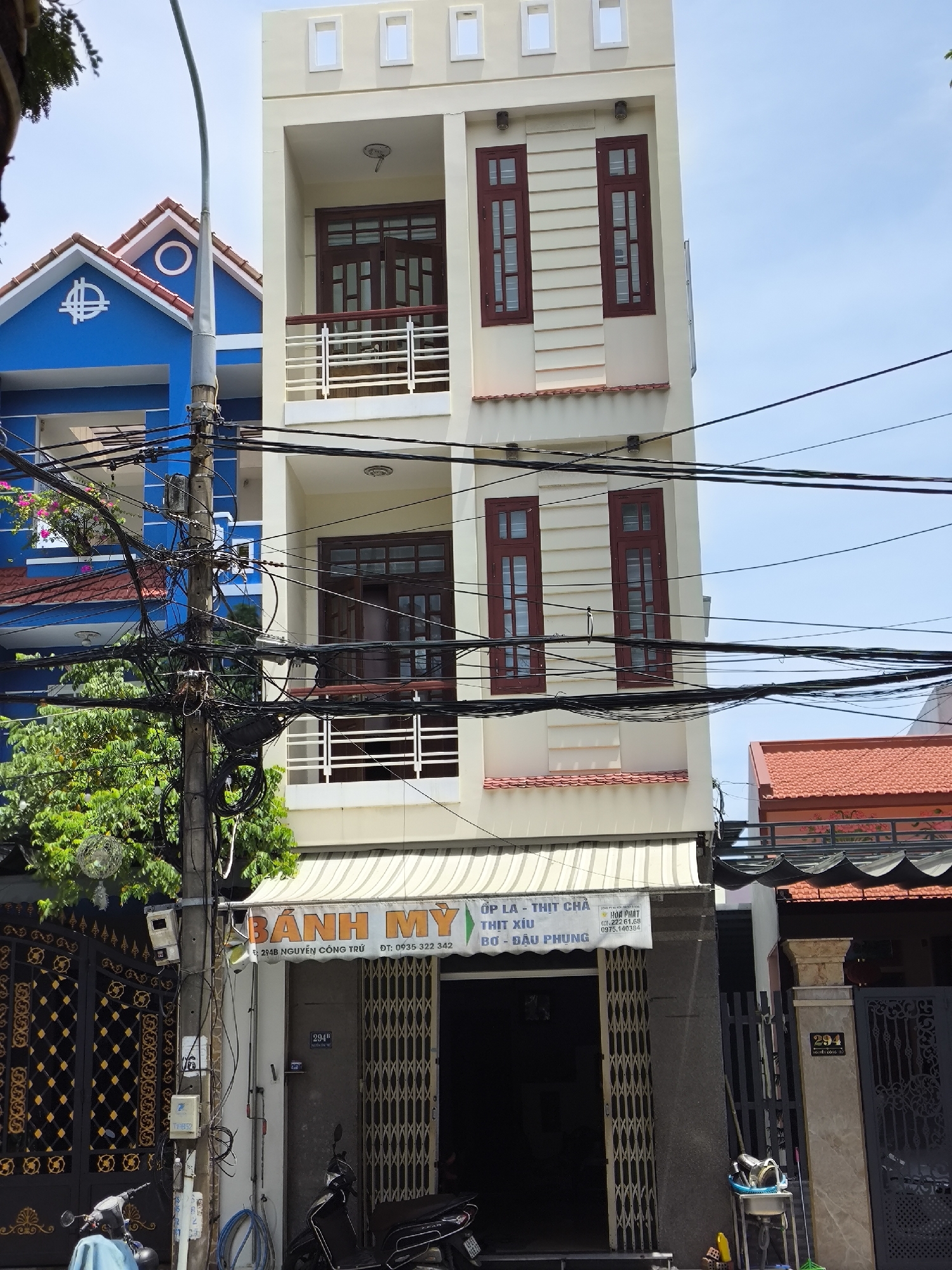This image shows a narrow, three-story building in Da Nang, Vietnam, with a storefront on the ground floor. The building is painted a light cream color and has dark red window frames and balconies with white railings. The storefront is covered by a retractable awning and has a sign that reads "BÁNH MỲ" (Vietnamese for baguette sandwich). Below this, smaller text indicates the types of fillings offered, such as "ỐP LA - THỊT CHẢ," "THỊT XÍU," and "BƠ - ĐẬU PHUNG." There is also a phone number listed. The entrance to the storefront is a metal security gate that is partially open, revealing the interior.
To the left of the building is a bright blue house with a red-tiled roof, and a utility pole with a dense tangle of electrical wires is positioned in front of both structures. Several motorcycles are parked on the sidewalk in front of the storefront. The street appears to be narrow, with other buildings and a red-tiled roof visible to the right. The sky is clear and blue.