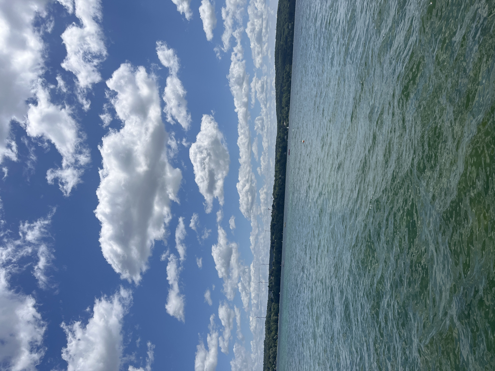 The image depicts a wide expanse of the Starnberger See in Starnberg, Germany, on a sunny day with a blue sky populated by numerous white, fluffy clouds. The water in the foreground exhibits a deep teal color with gentle ripples and waves, suggesting a light breeze. In the distance, a treed shoreline forms the horizon, dotted with a few buildings and several wind turbines, indicating a developed area. A few small boats and buoys are visible on the water, implying recreational activities. The overall scene is one of natural beauty and a pleasant atmosphere.